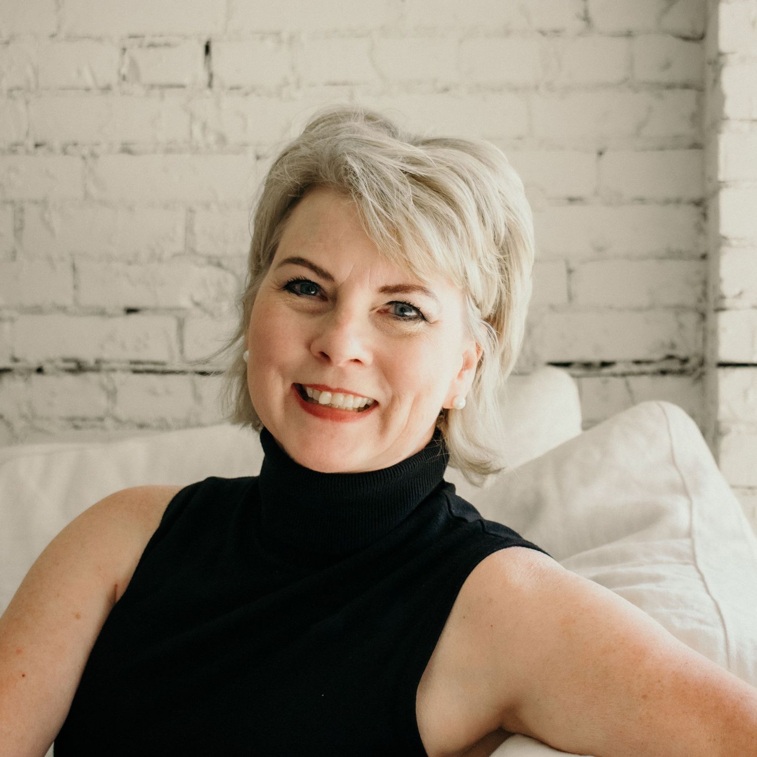 Professional headshot of a woman in black sleeveless top against white brick wall background.