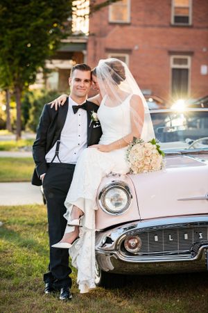 Bride and groom pose beside a vintage pink Oldsmobile classic car at golden hour.
