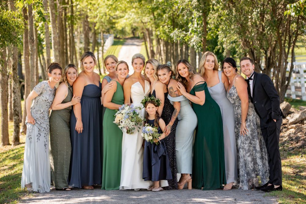Bride poses with bridesmaids in navy and green gowns among tree-lined path.