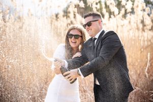 Bride and groom celebrate with champagne spray outdoors among golden tall grasses.
