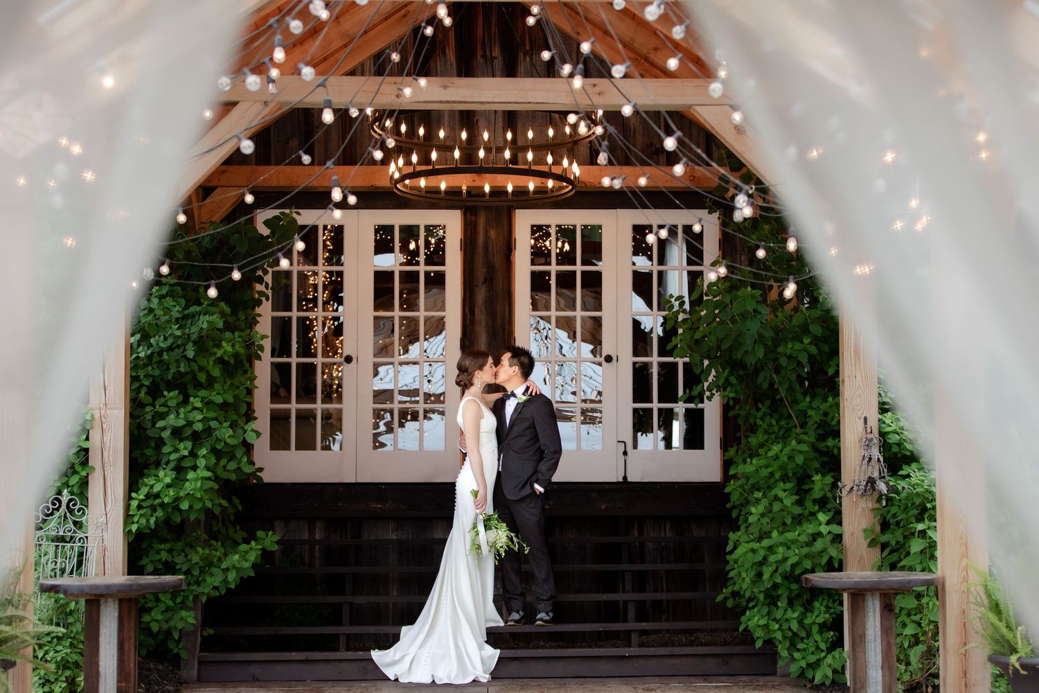 Bride and groom sharing a kiss under string lights at a rustic barn wedding venue entrance.
