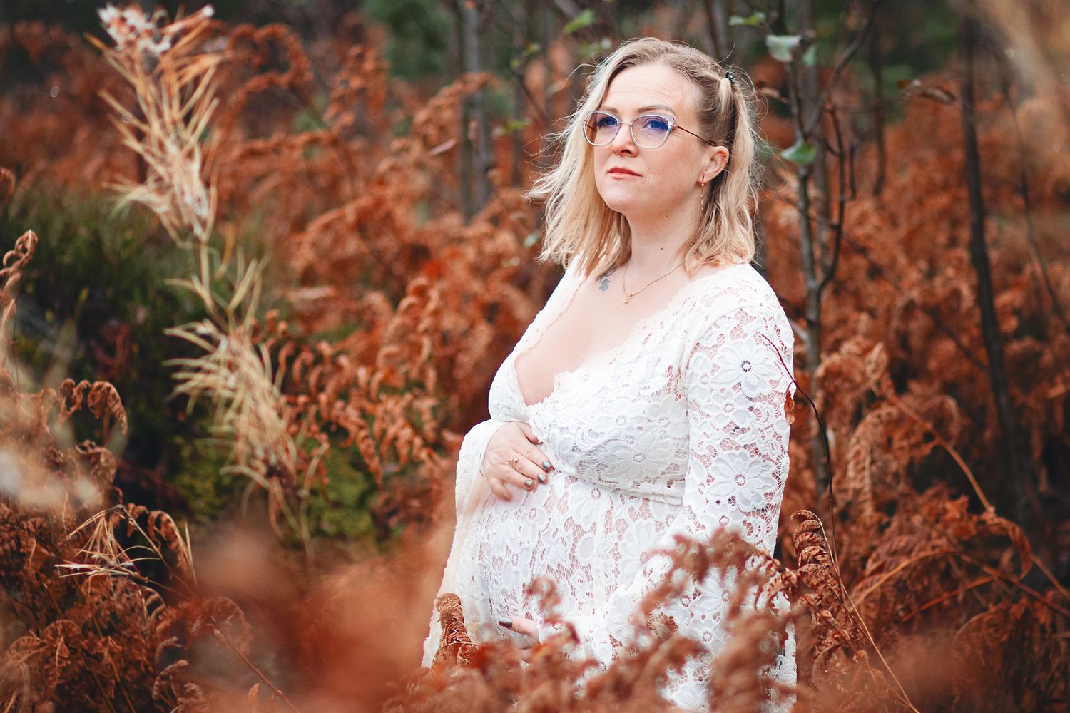 A woman in white lace poses among rustic autumn ferns during a maternity photoshoot.