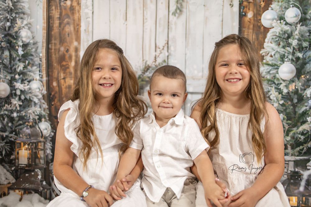 Three children in white outfits smiling together in a festive Christmas studio backdrop.