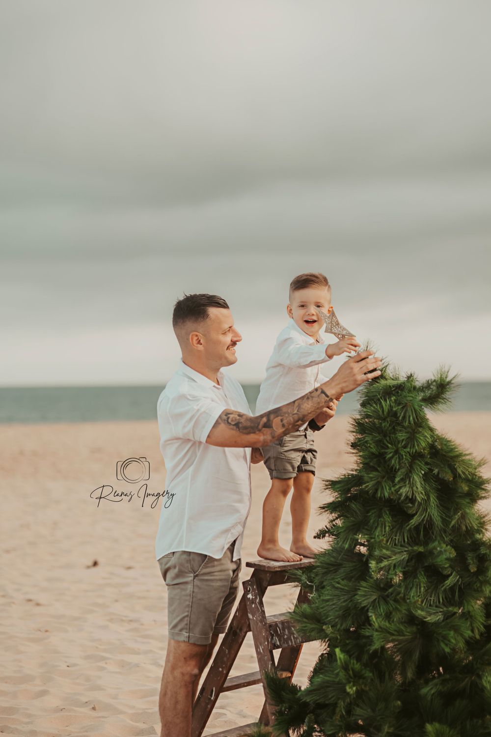 Smiling father lifting laughing toddler son beside a Christmas tree on the beach.