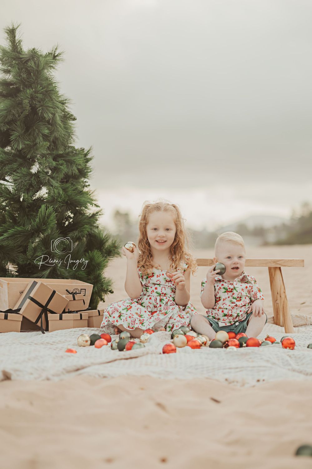 Two sisters in matching floral dresses sitting by a Christmas tree on sandy beach.