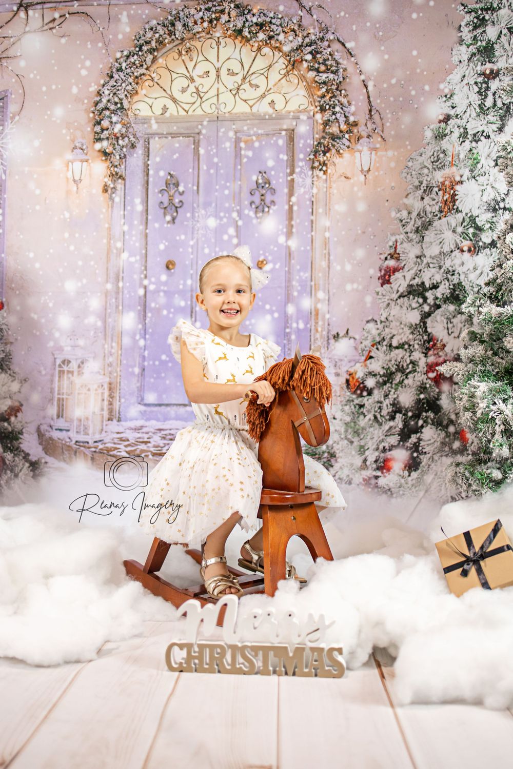 Young girl in white dress riding a rocking horse in a magical Christmas studio setting.