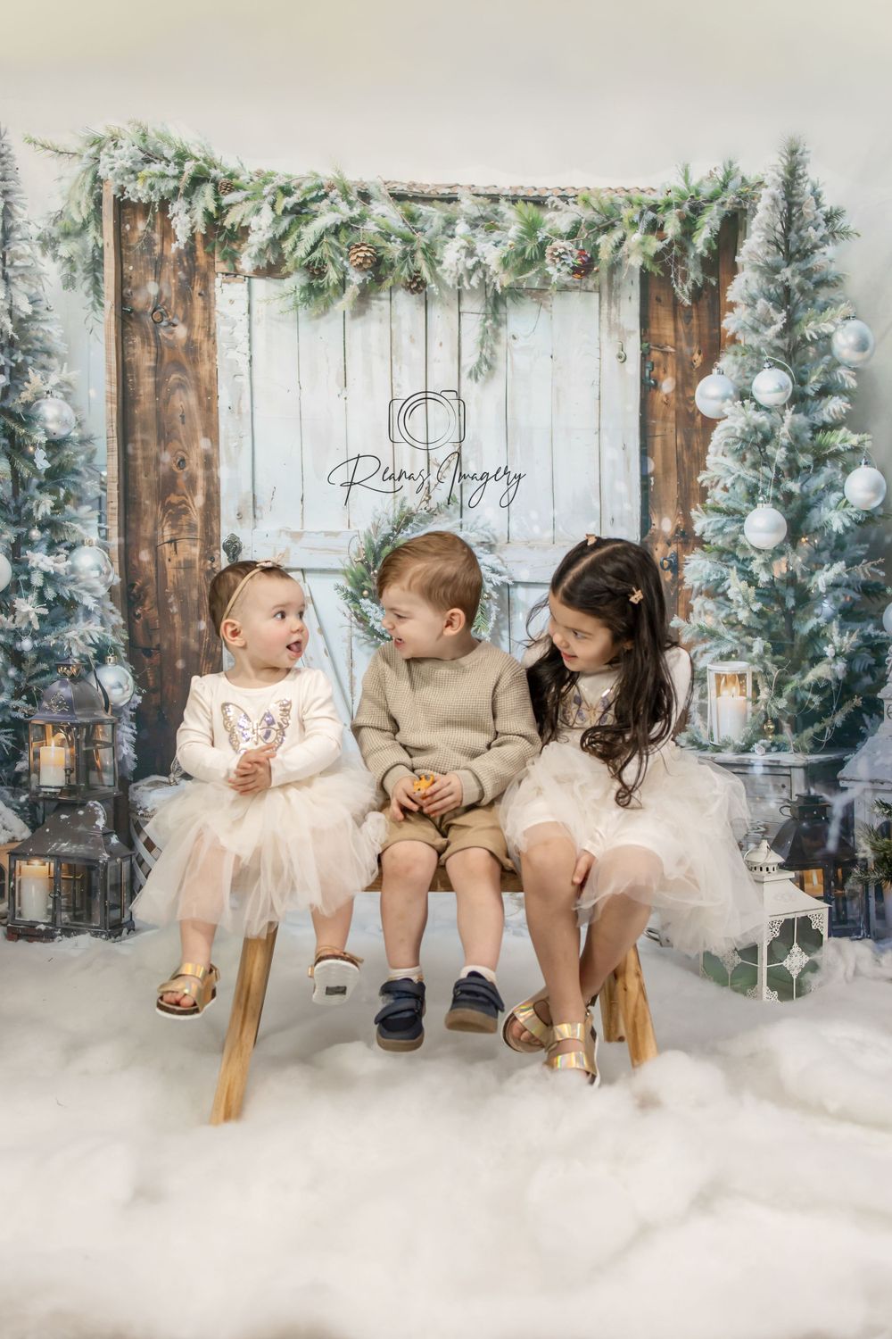 Three young siblings in white outfits sitting together in a winter Christmas studio set.
