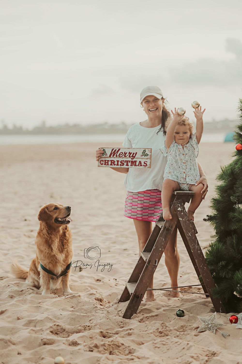 Mother and daughter holding Merry Christmas sign with golden retriever on sunny beach.