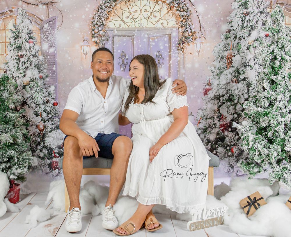 Couple smiling together in a festive Christmas photo studio with snowy trees and holiday decorations.
