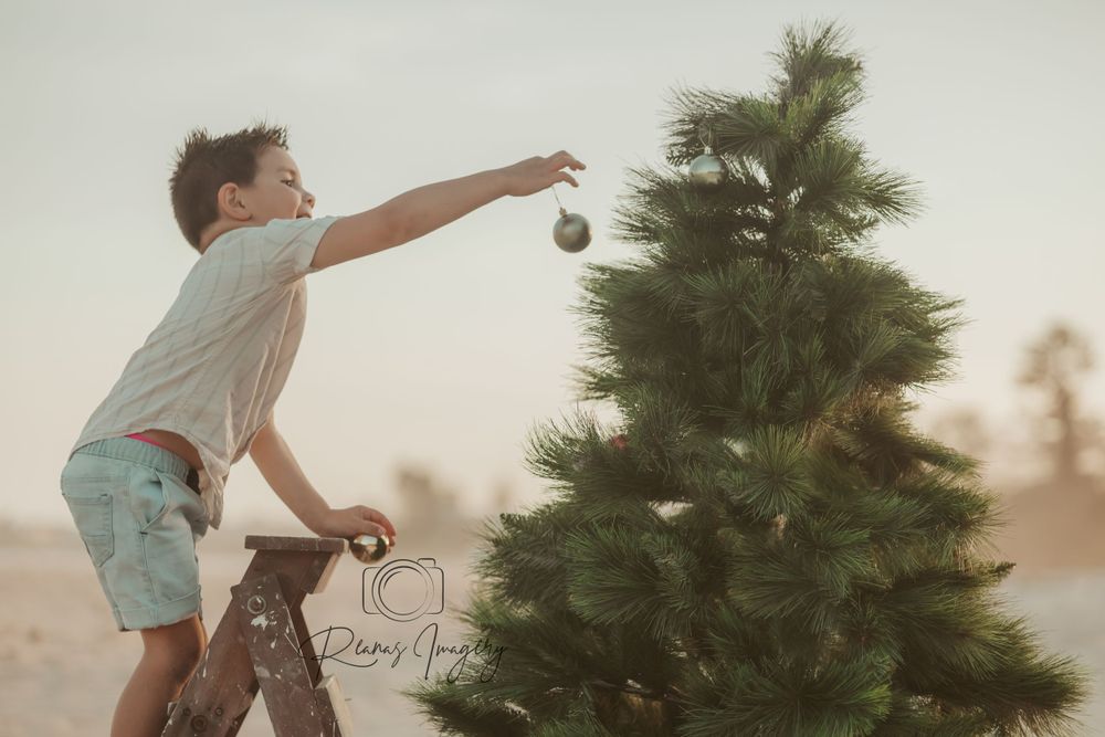 Young boy reaches to place ornament on top of a Christmas tree outdoors at sunset.