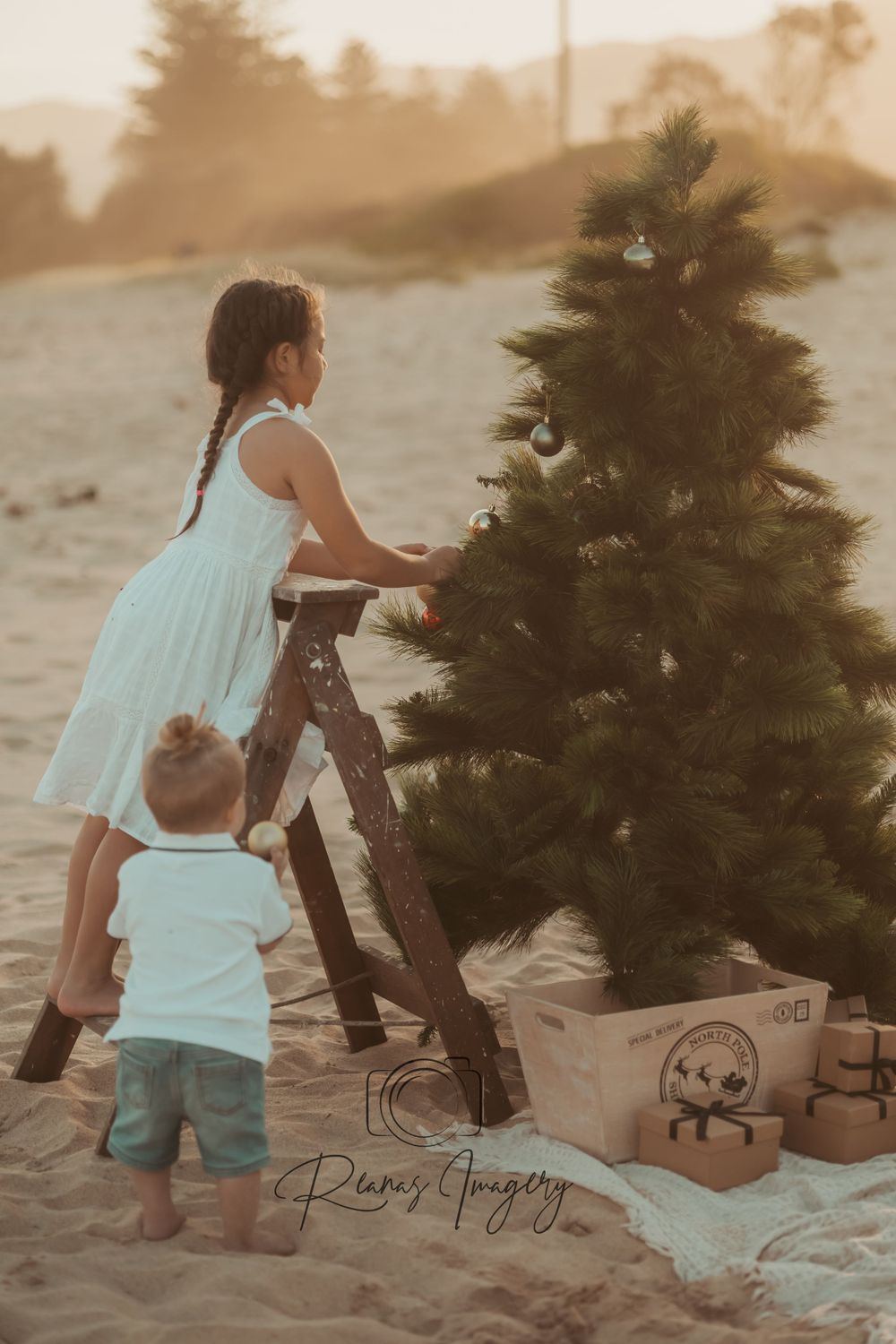 Two children decorate a Christmas tree on a sandy beach at golden hour.