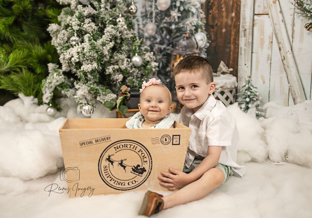 Baby and young boy sit together in a wooden Santa delivery crate Christmas photo prop.