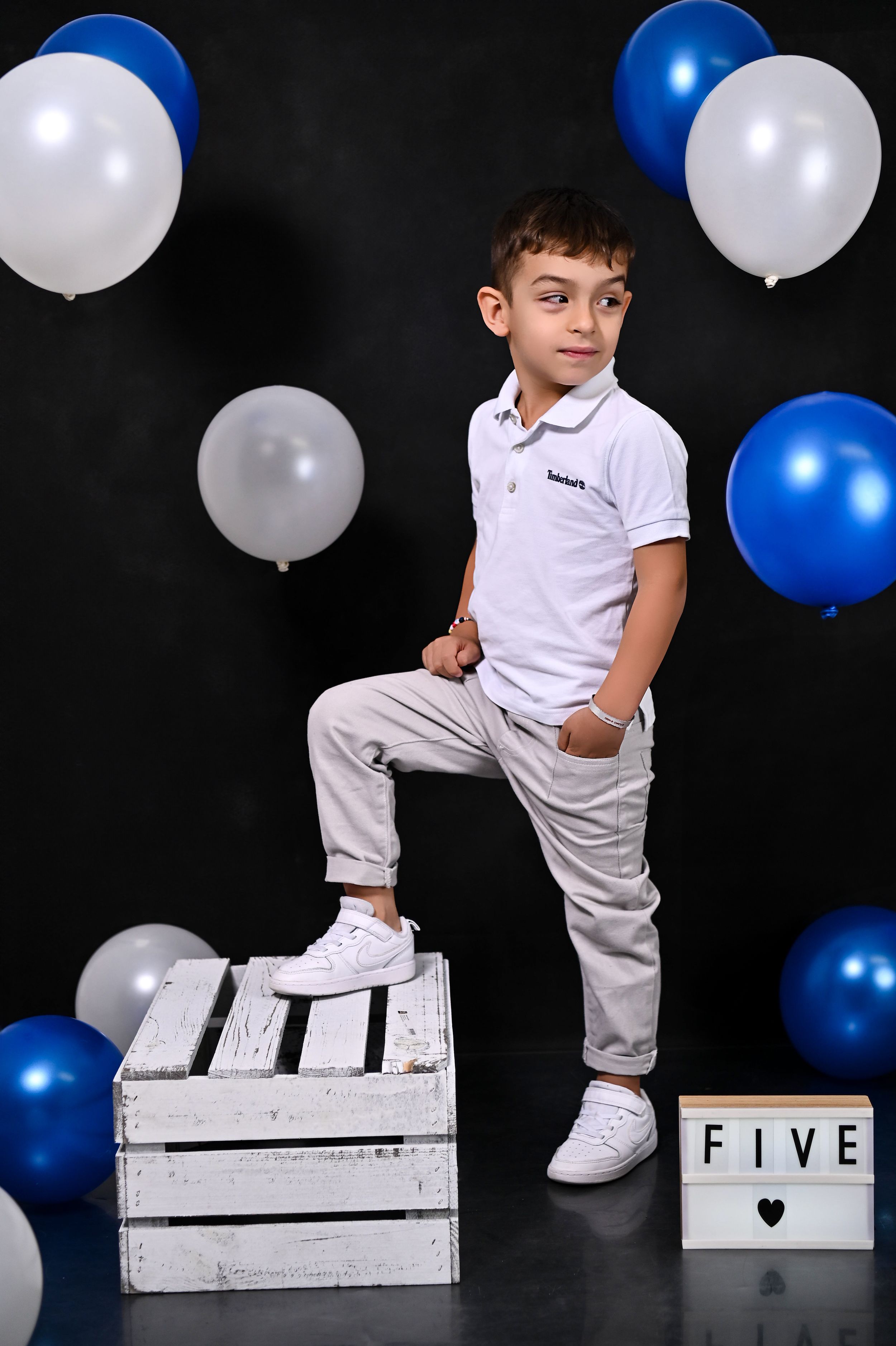 Child in white outfit poses on wooden crates with blue and white balloons against black background.