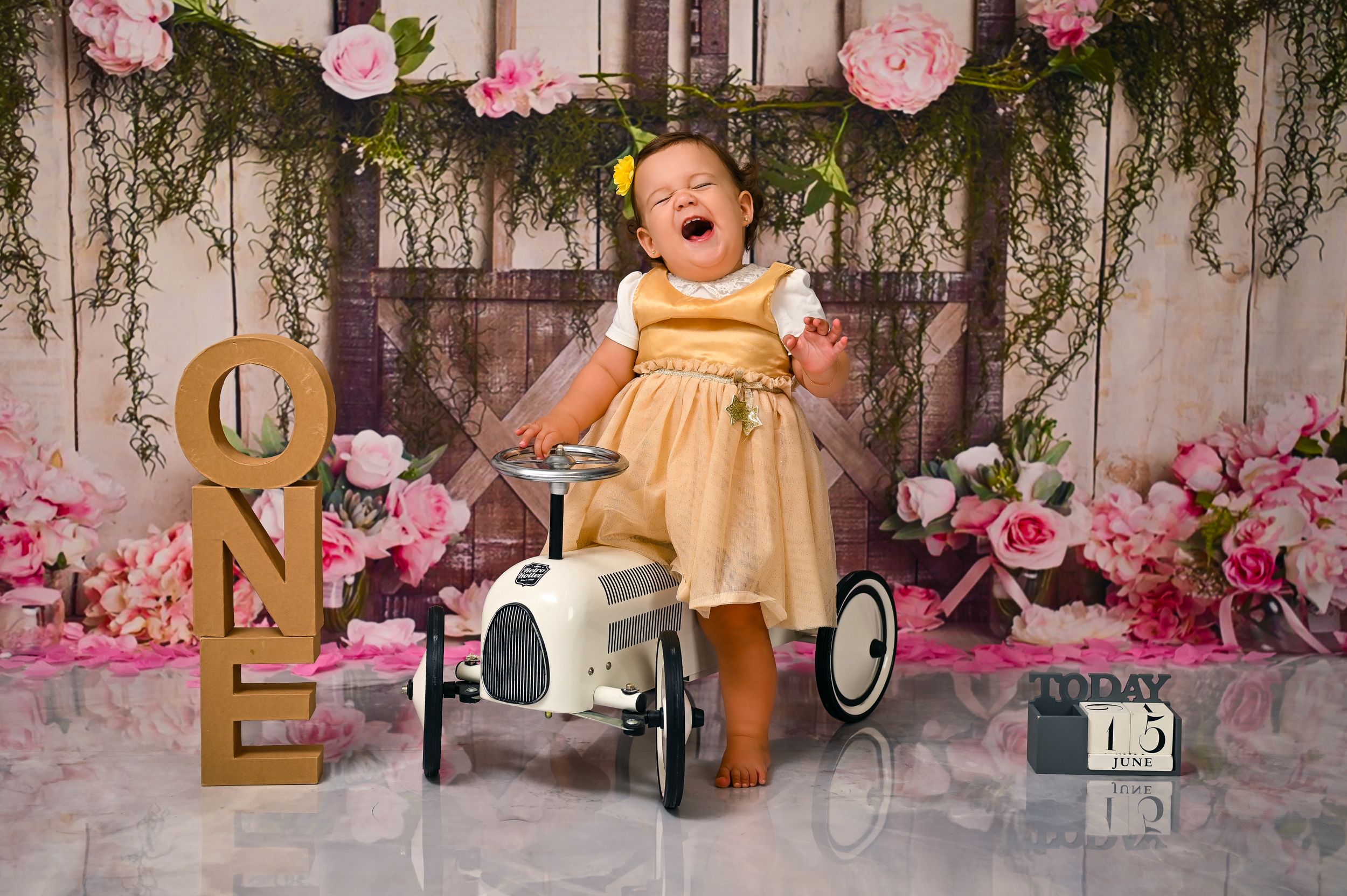 Baby in yellow dress poses with vintage pedal car for first birthday photoshoot.