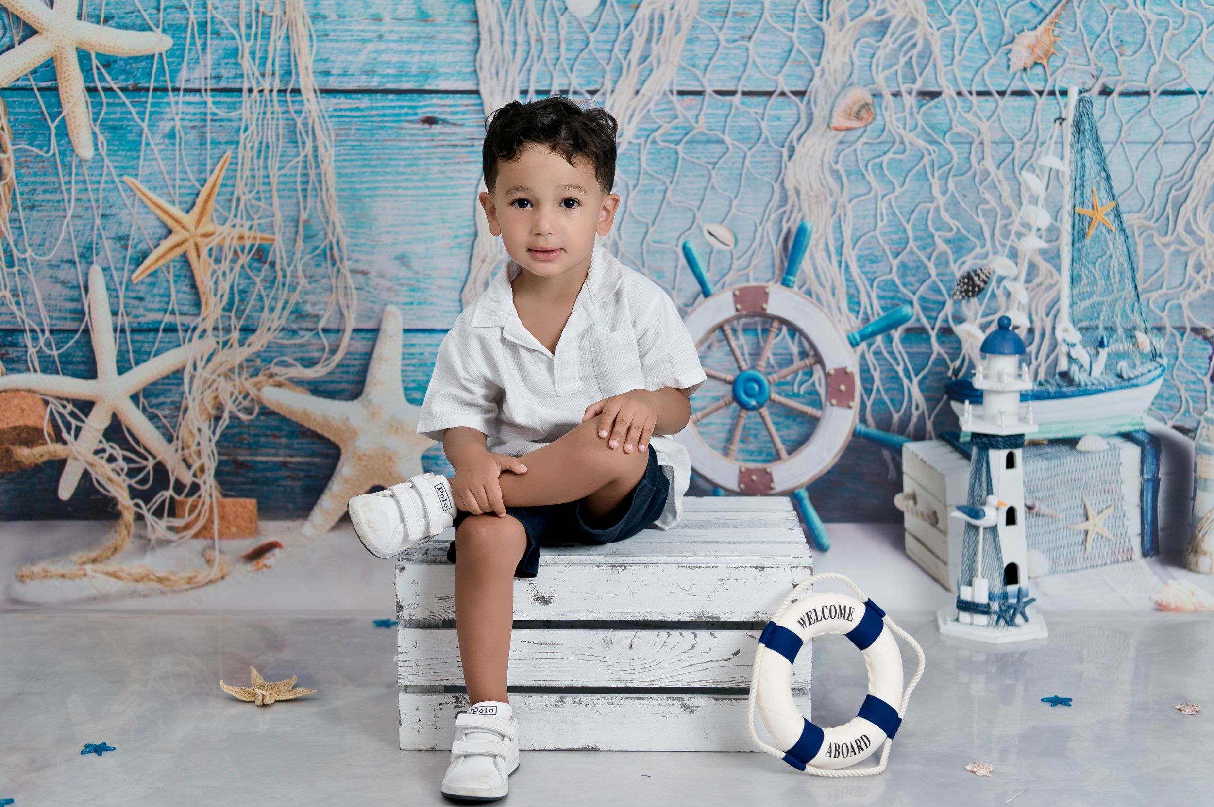 Nautical themed photo setup with white boat wheel, starfish decorations, and lighthouse props against blue backdrop.