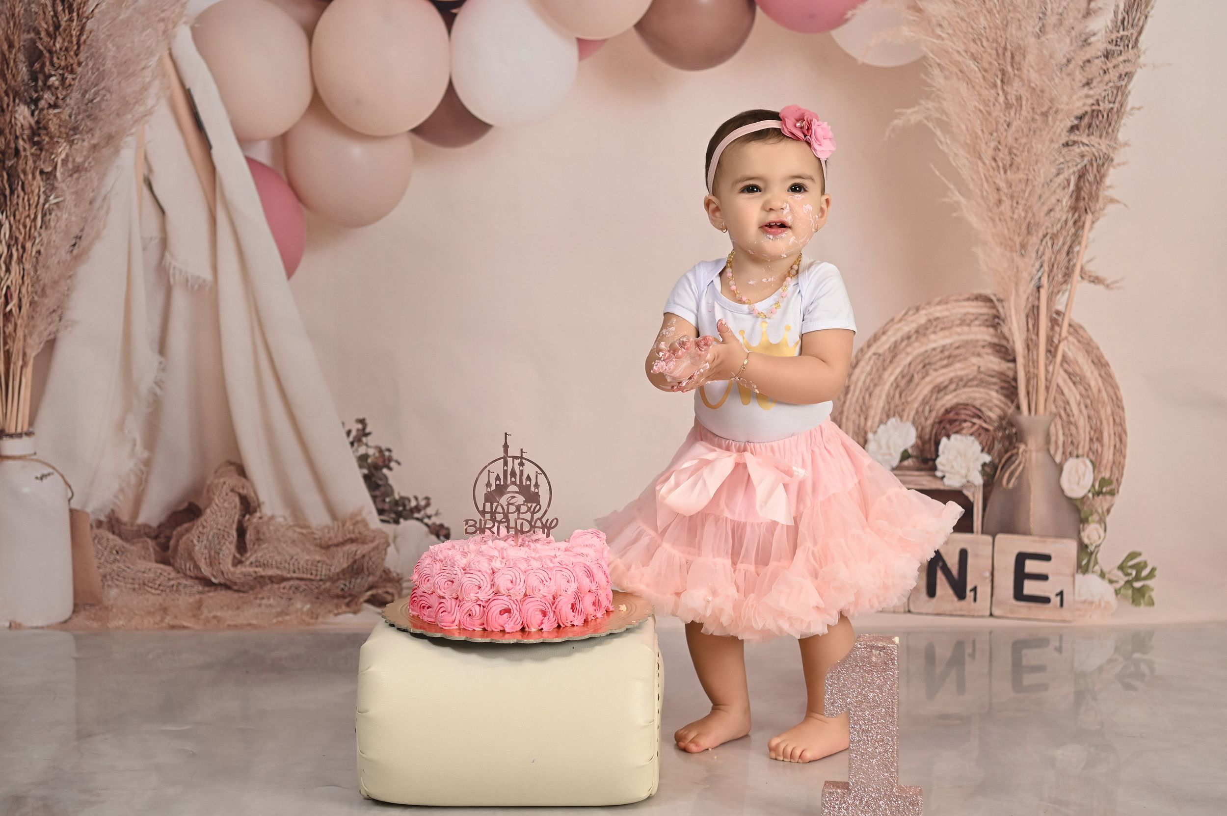 Pink birthday setup with tulle skirt, balloon arch, and pink cake on cream ottoman.