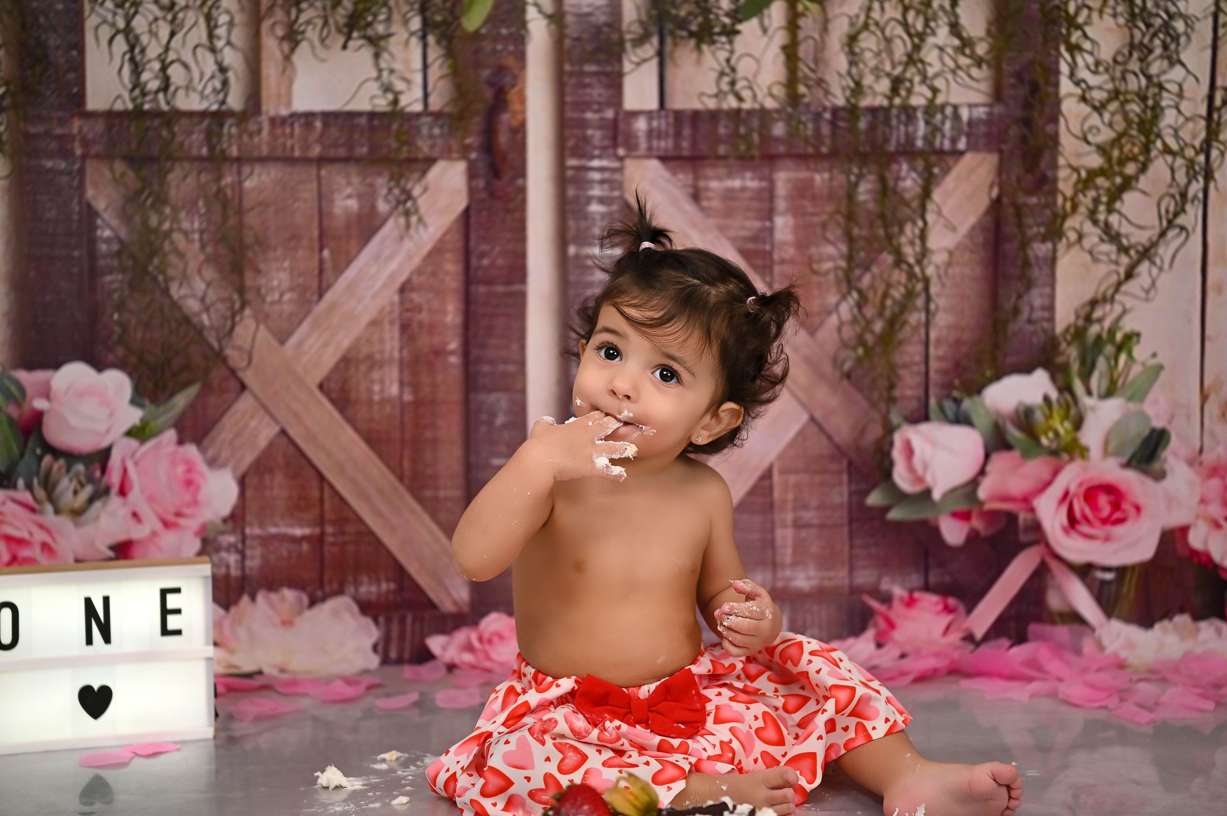 Baby in red tutu smiles during cake smash photo session with pink floral decorations.