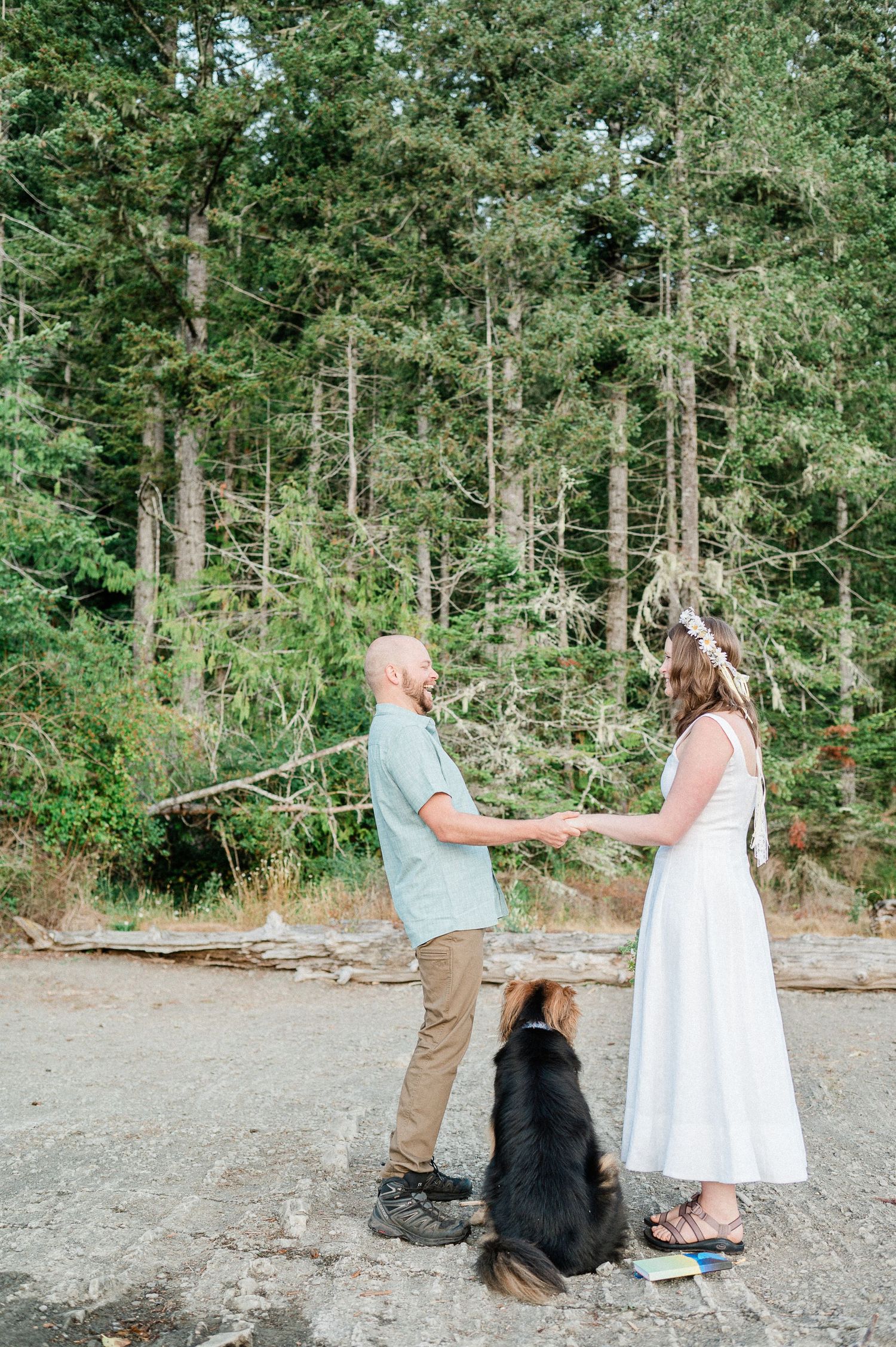A couple stands together on a forest path with their black dog sitting between them on a sunny day.
