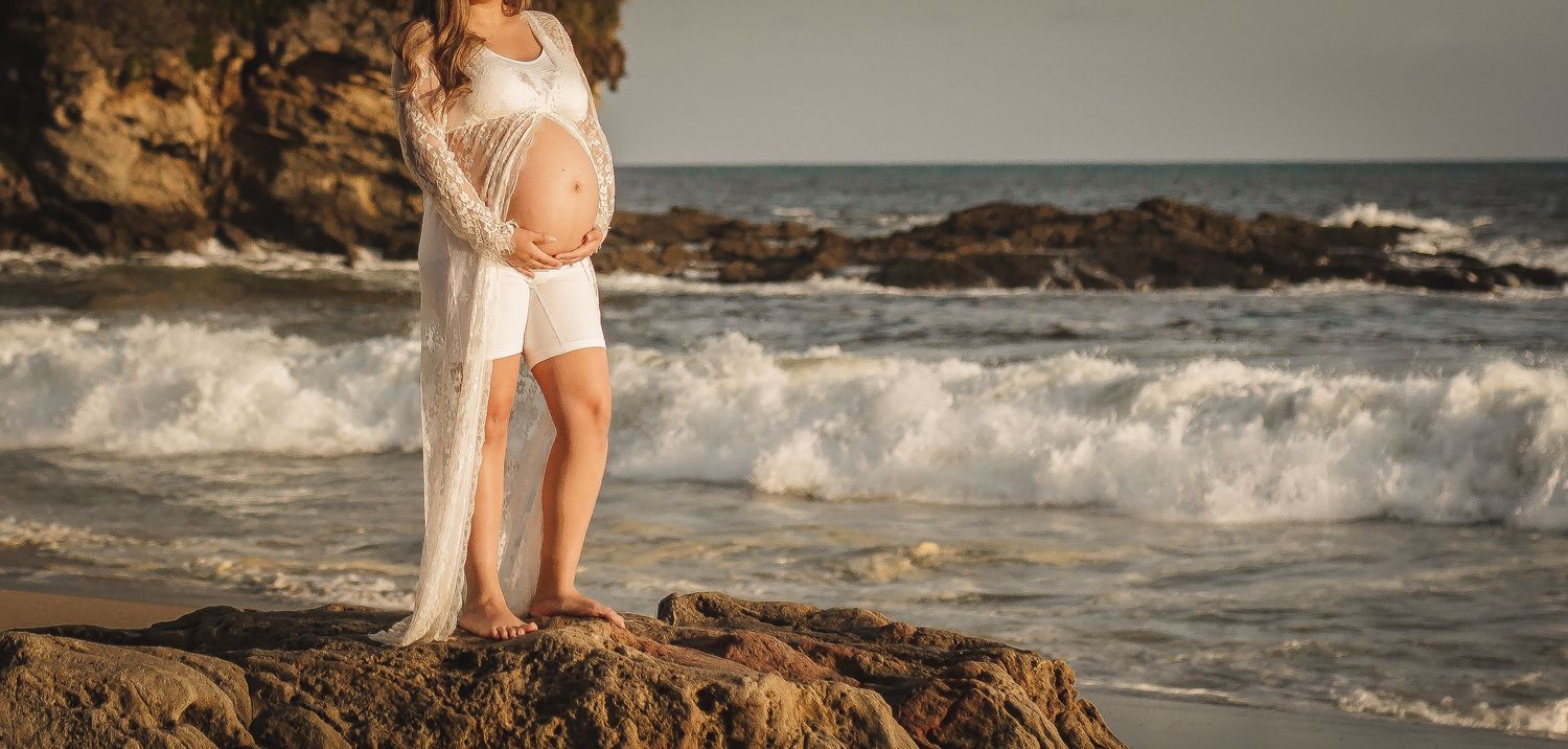 Artistic maternity photo with waves crashing against coastal rocks at sunset.
