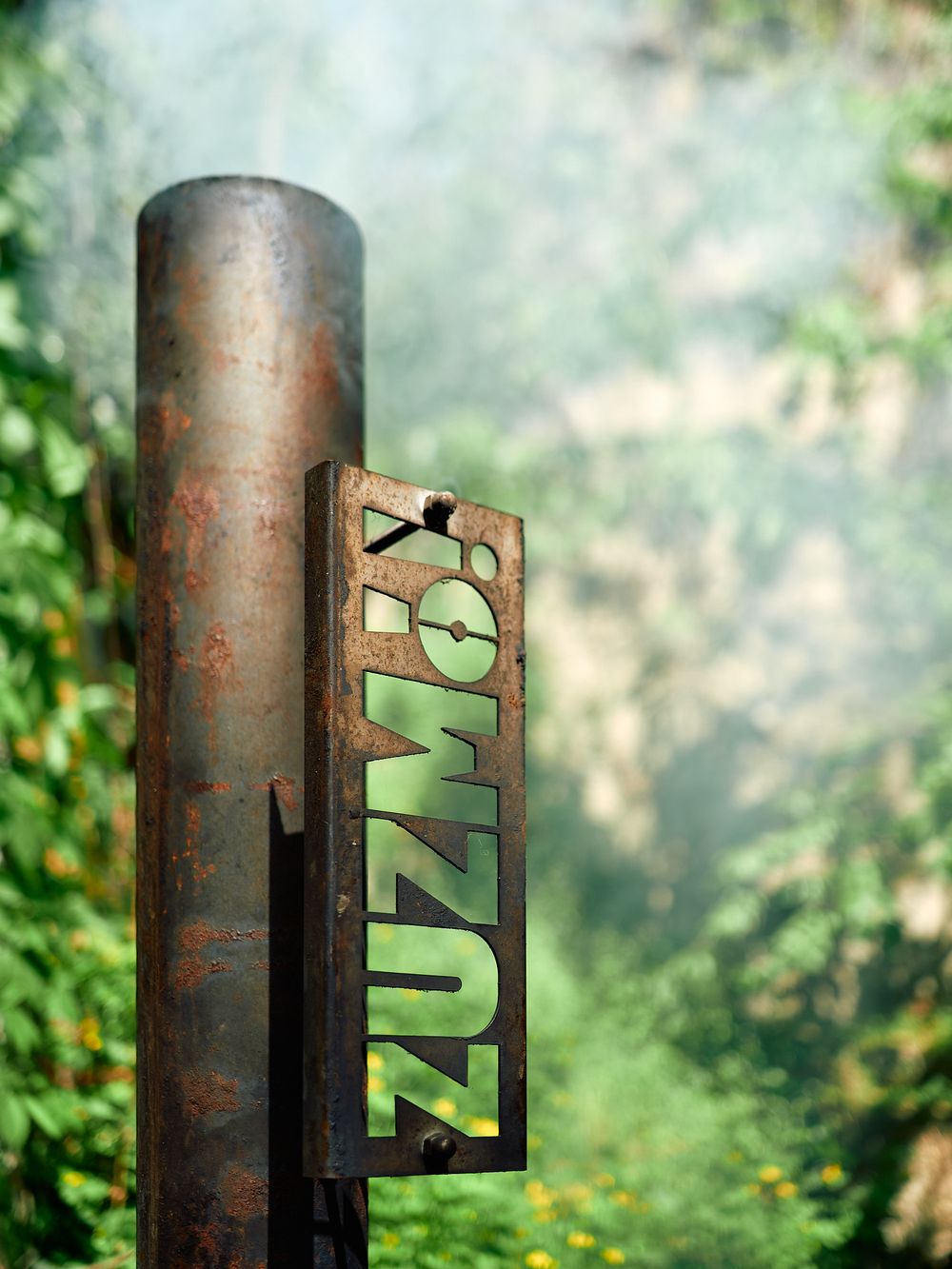 Rustic metal pole with green 'Zuzmó' text cutout against blurred natural background.