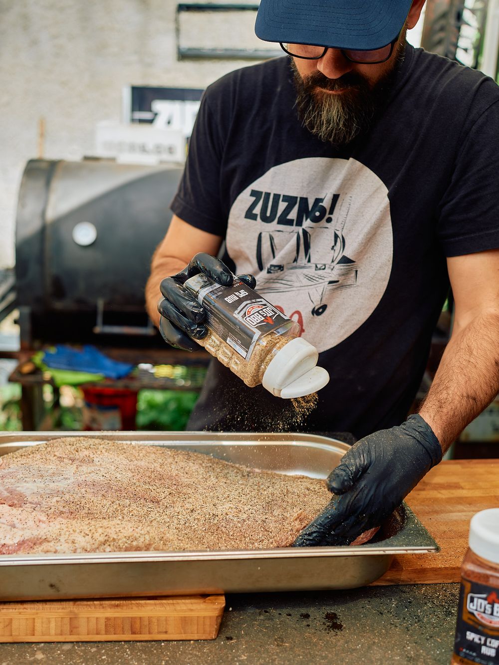 Seasoning meat with spices from glass shaker at outdoor cooking station.