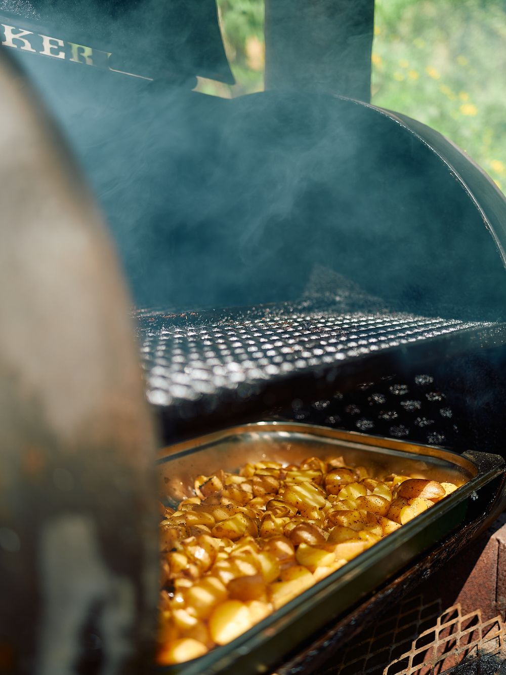 Close-up of smoking barbecue grill with golden macaroni and cheese dish cooking inside.