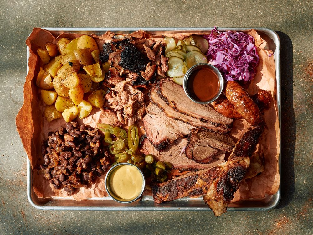 BBQ platter with smoked meats, potatoes, purple slaw and two dipping sauces on metal tray.