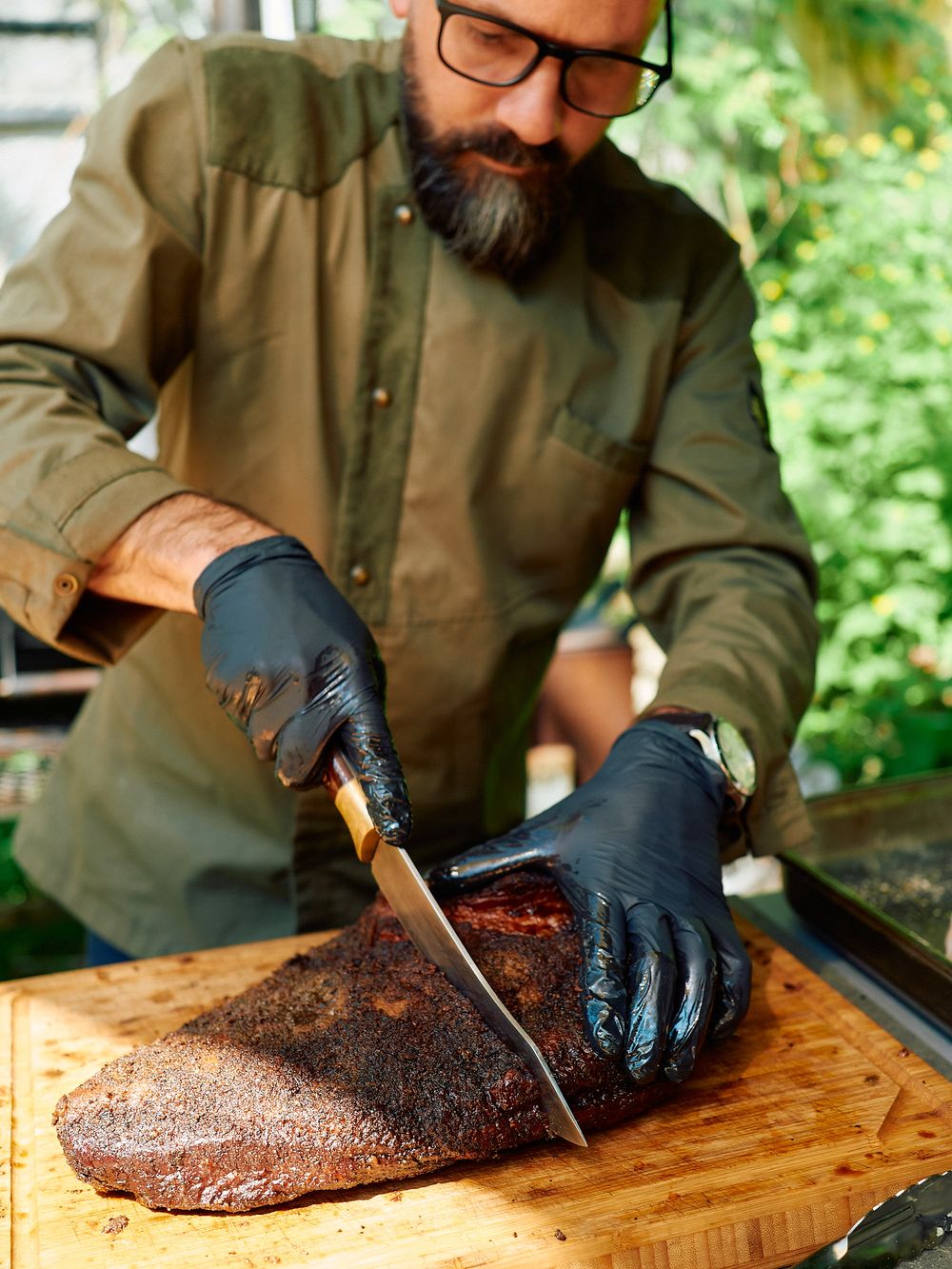 Cook in olive jacket slicing smoked brisket on wooden cutting board outdoors.