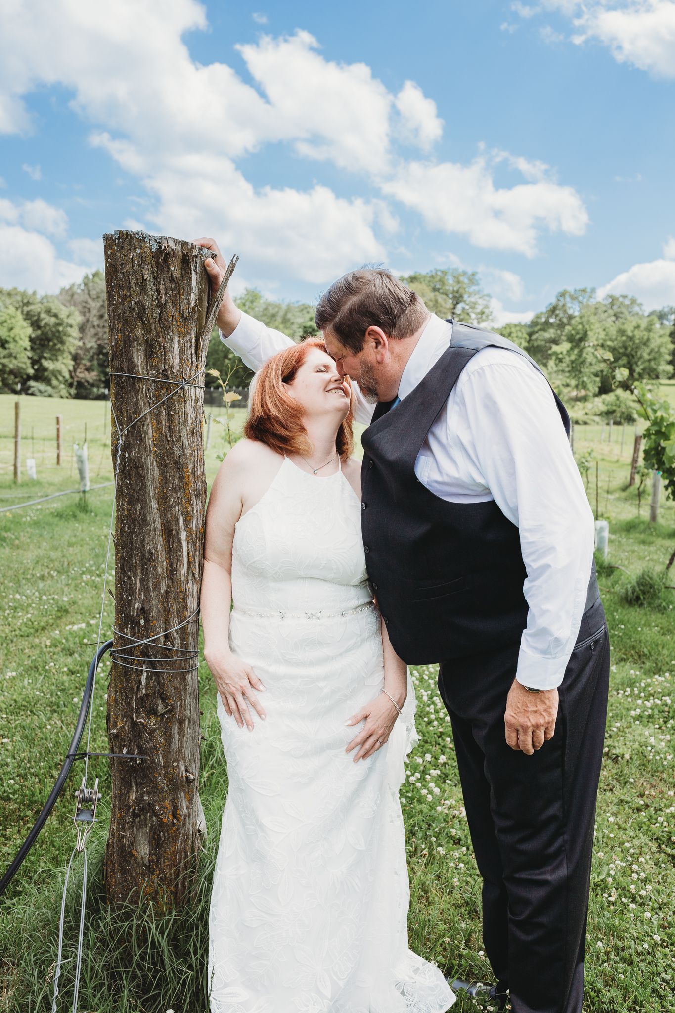 Newlywed couple shares an intimate moment by a rustic wooden fence post in a sunny countryside setting.