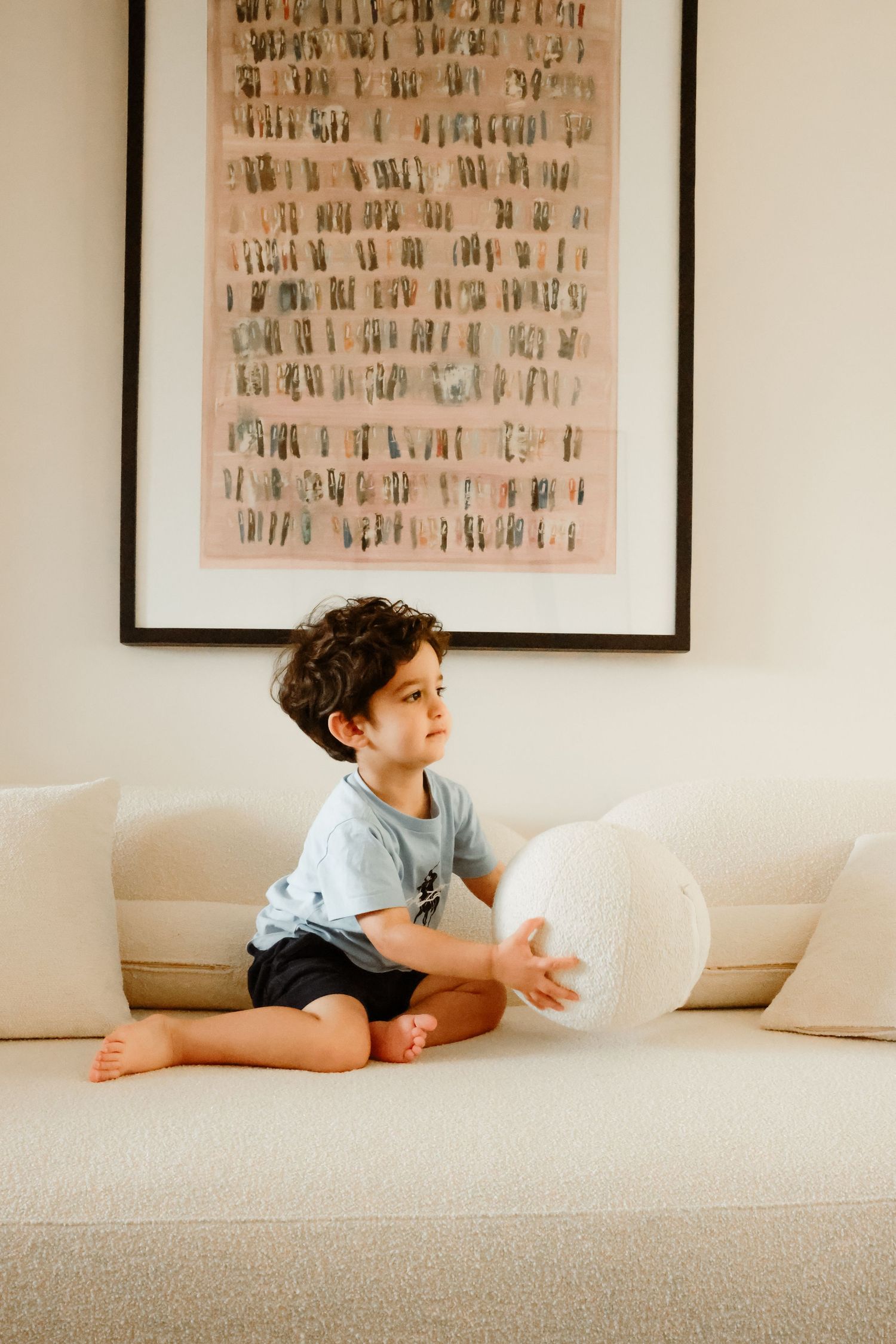 A child sits cross-legged on a beige couch with a white balloon under abstract wall art.