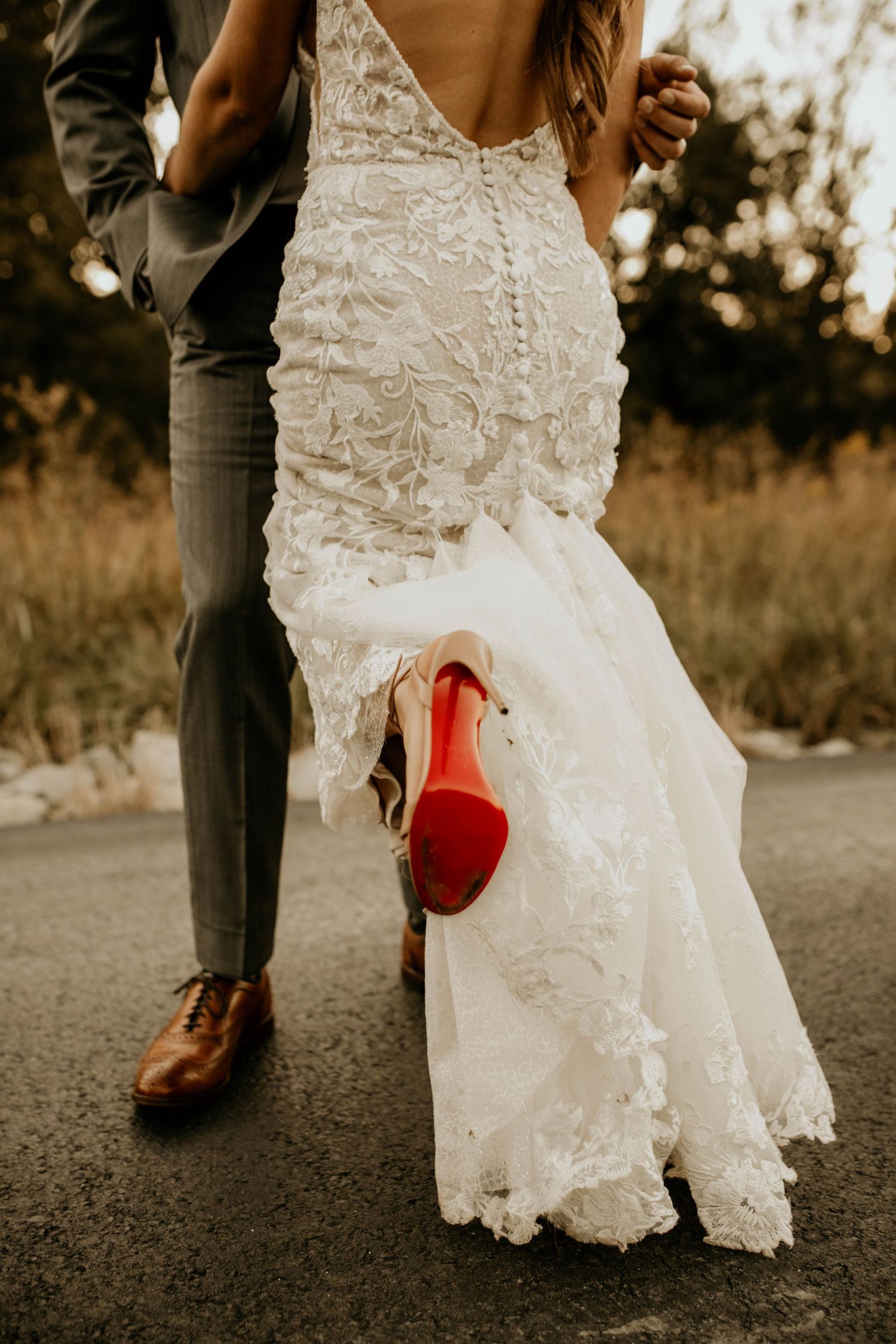 Bride in lace mermaid gown with red-sole heels walks hand-in-hand with groom on outdoor path.