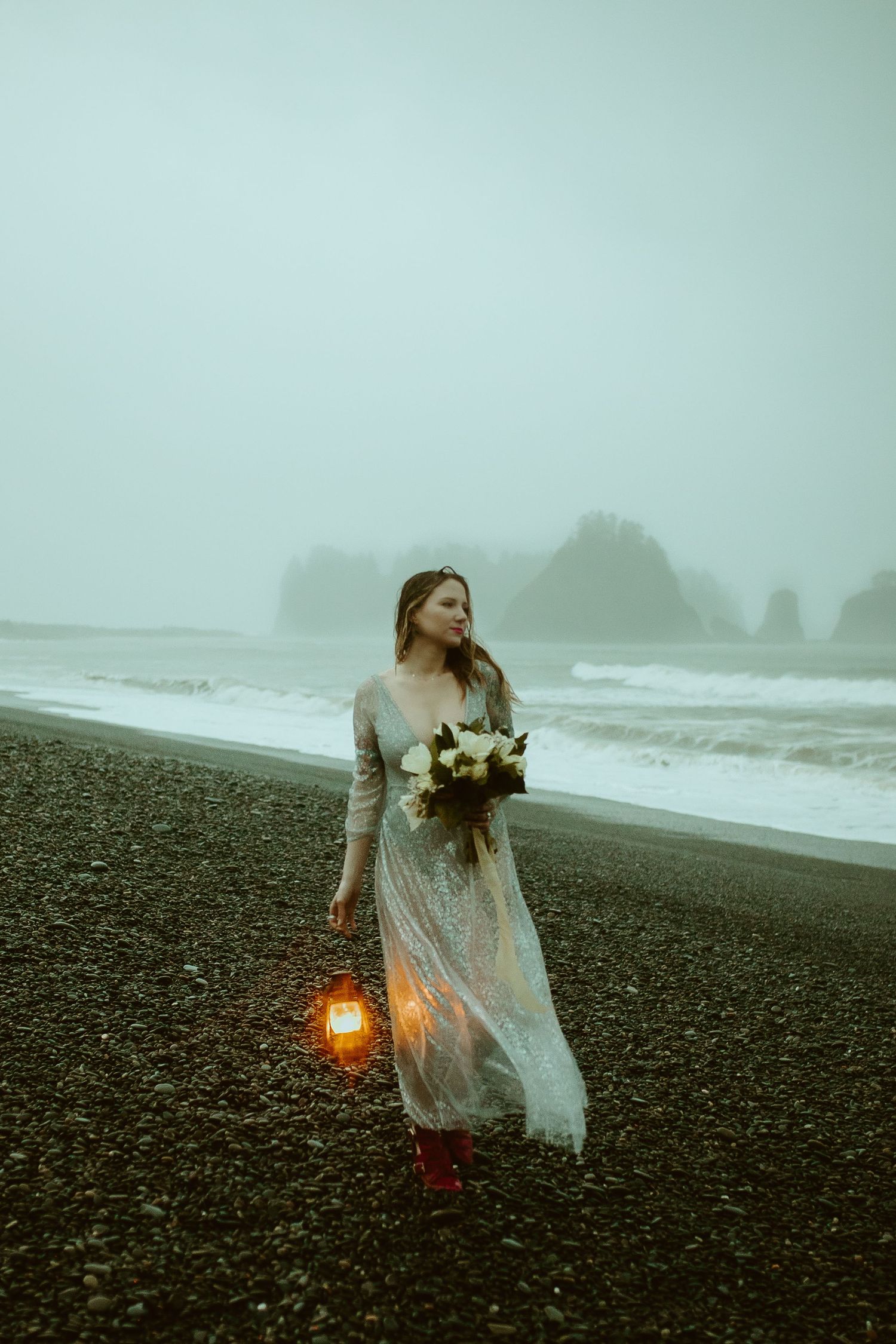 Bride in lace gown holding lantern and bouquet walks on misty dark beach.
