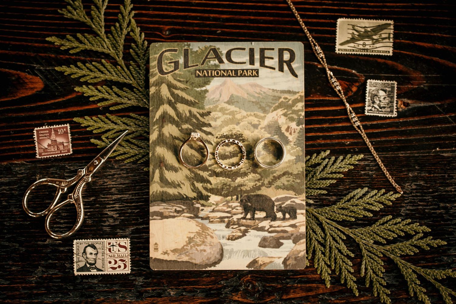 Glacier National Park journal with wedding rings rests among vintage stamps and scissors on dark wood.