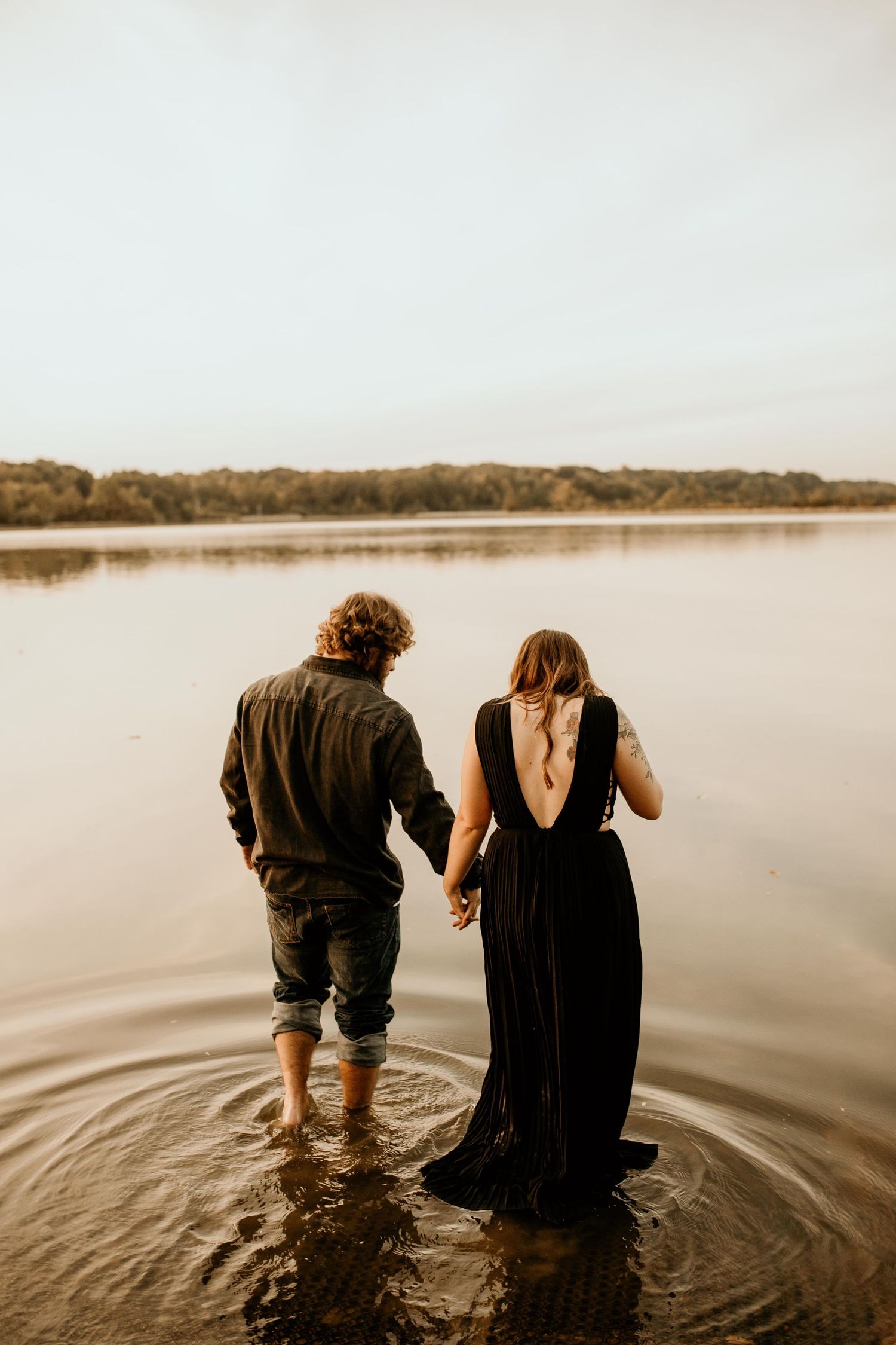 Couple holding hands while wading in calm lake water at sunset, wearing casual black outfits.