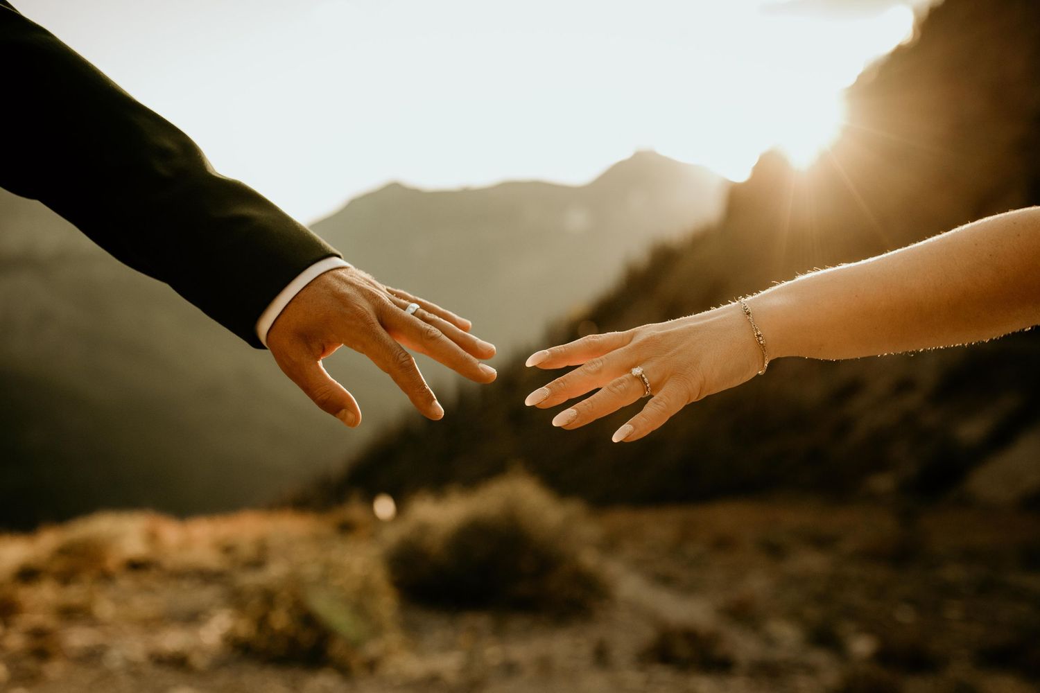 Bride and groom reach toward each other with golden mountain sunset glowing behind their hands.