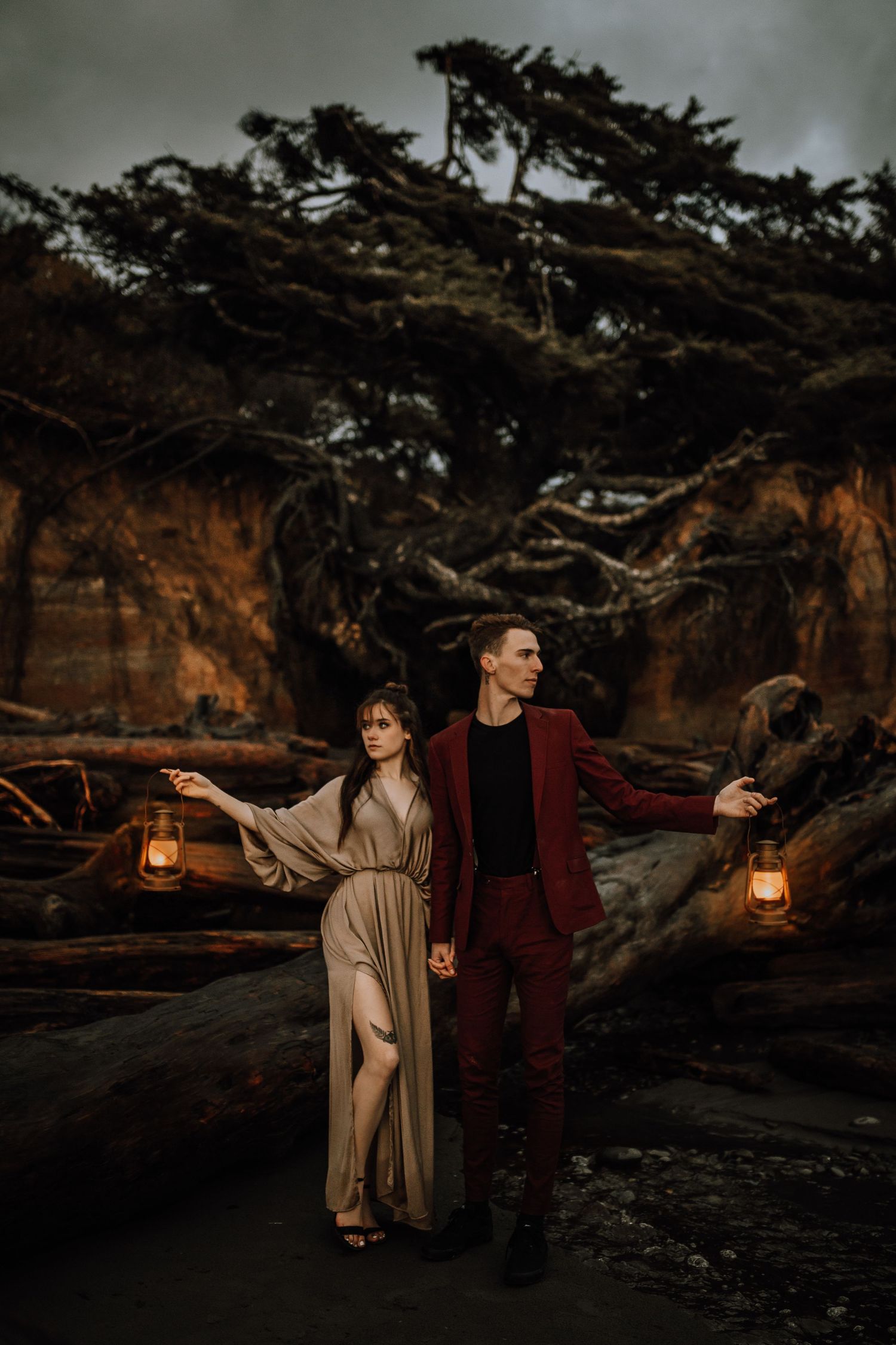 Elegant couple holding vintage lanterns on a dramatic driftwood beach at dusk.