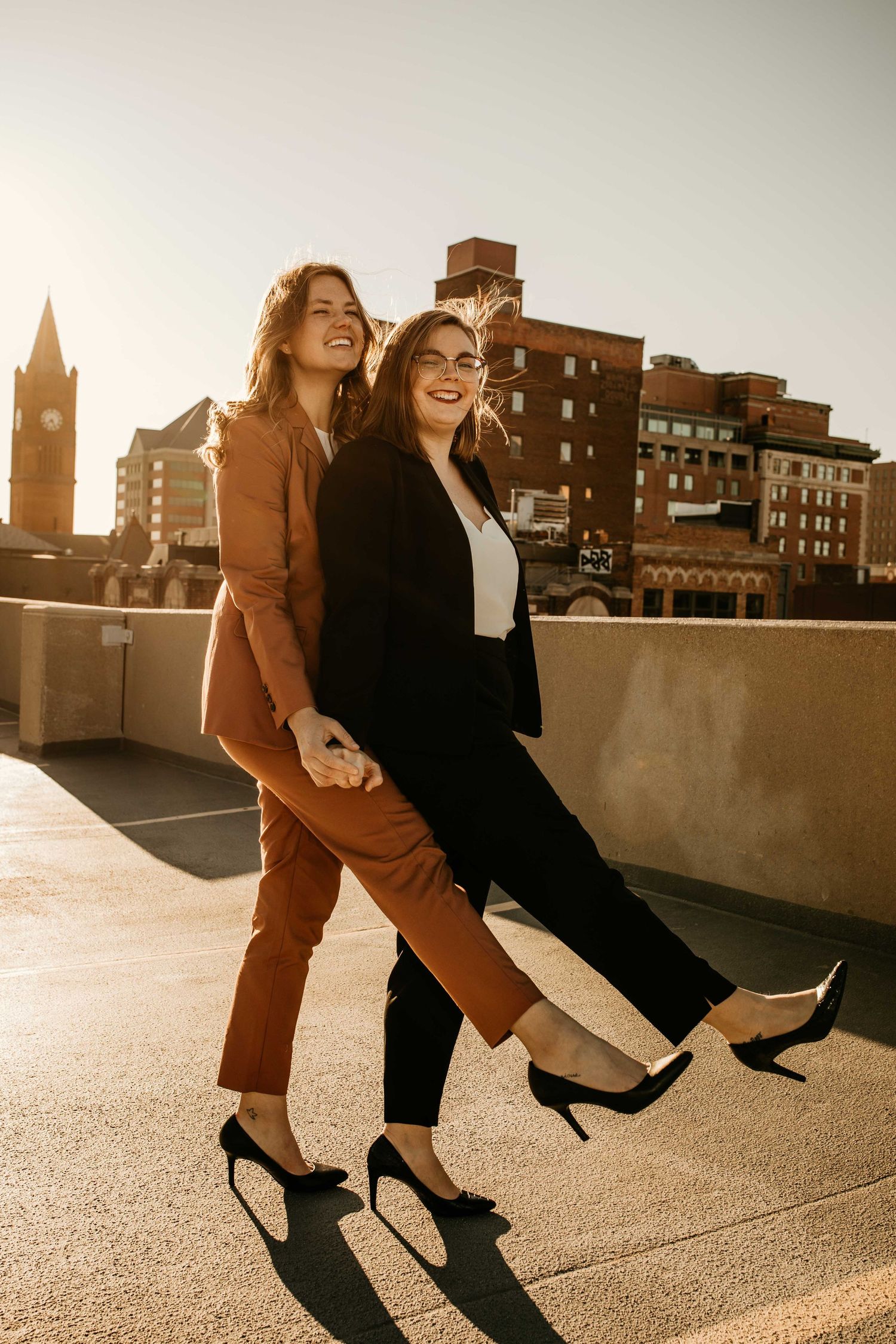 Two professional women in stylish suits laugh and walk together on a rooftop at golden hour.