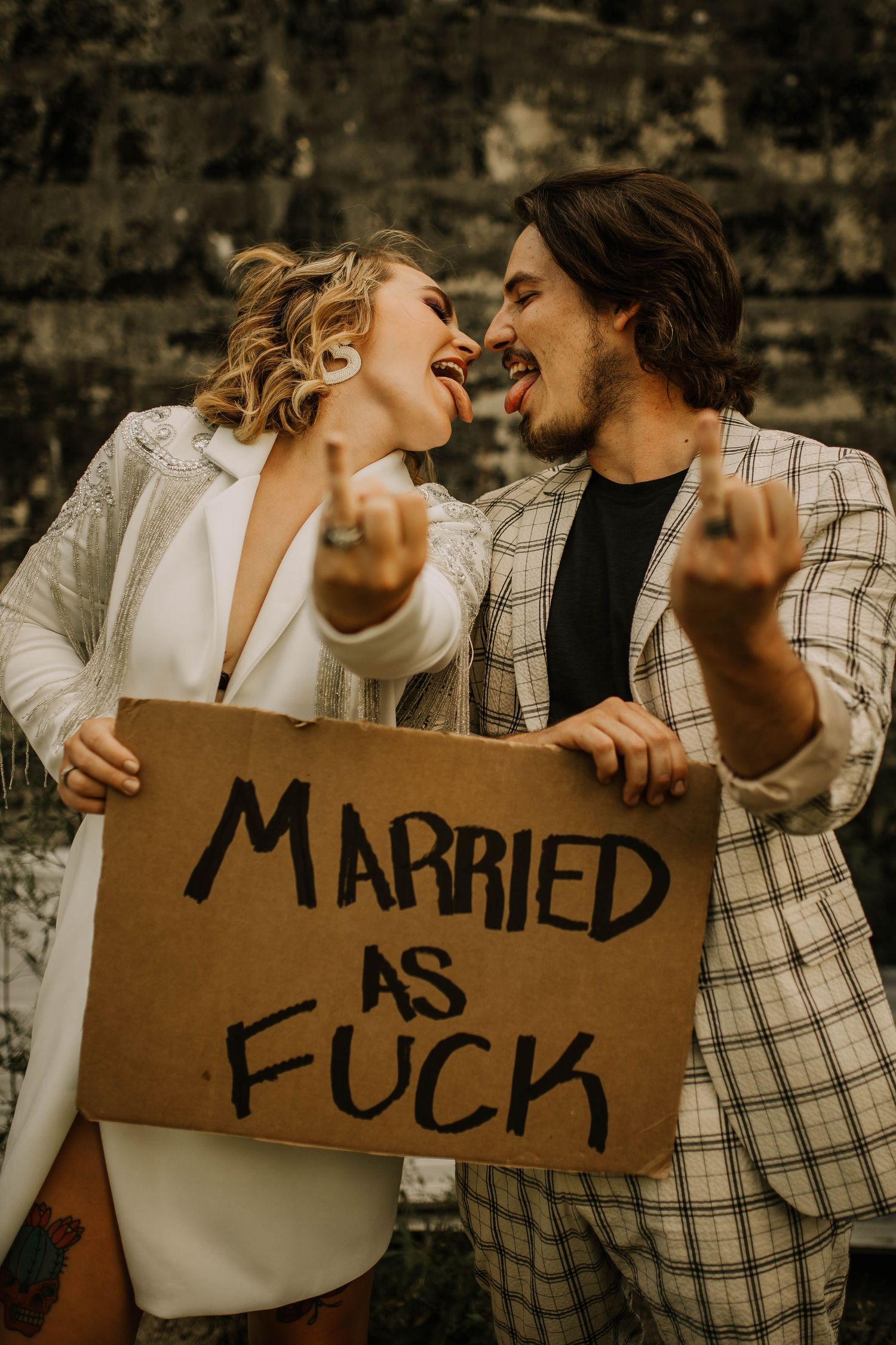 Laughing newlywed couple holds a cardboard sign reading Married As F*** at a fun rebellious wedding.