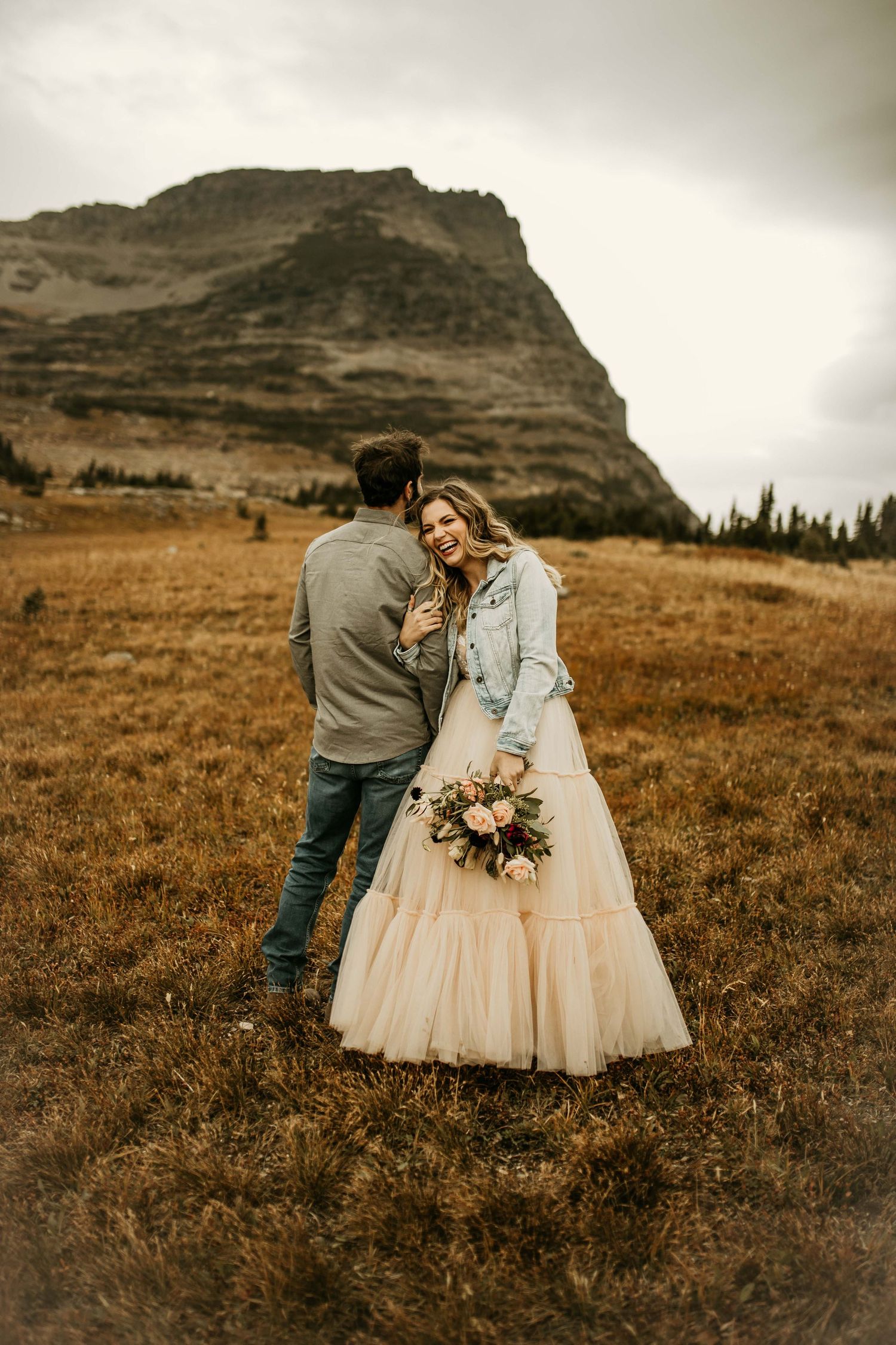 Bride in tulle gown and denim jacket holds a bouquet as groom embraces her in a dramatic mountain meadow.