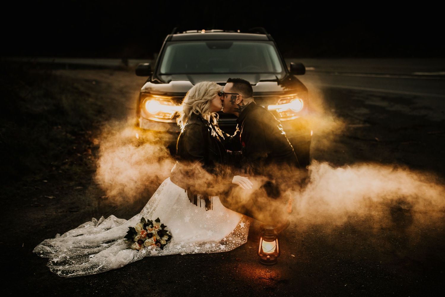 Bride and groom kissing in front of truck headlights with smoke and a lantern at night.