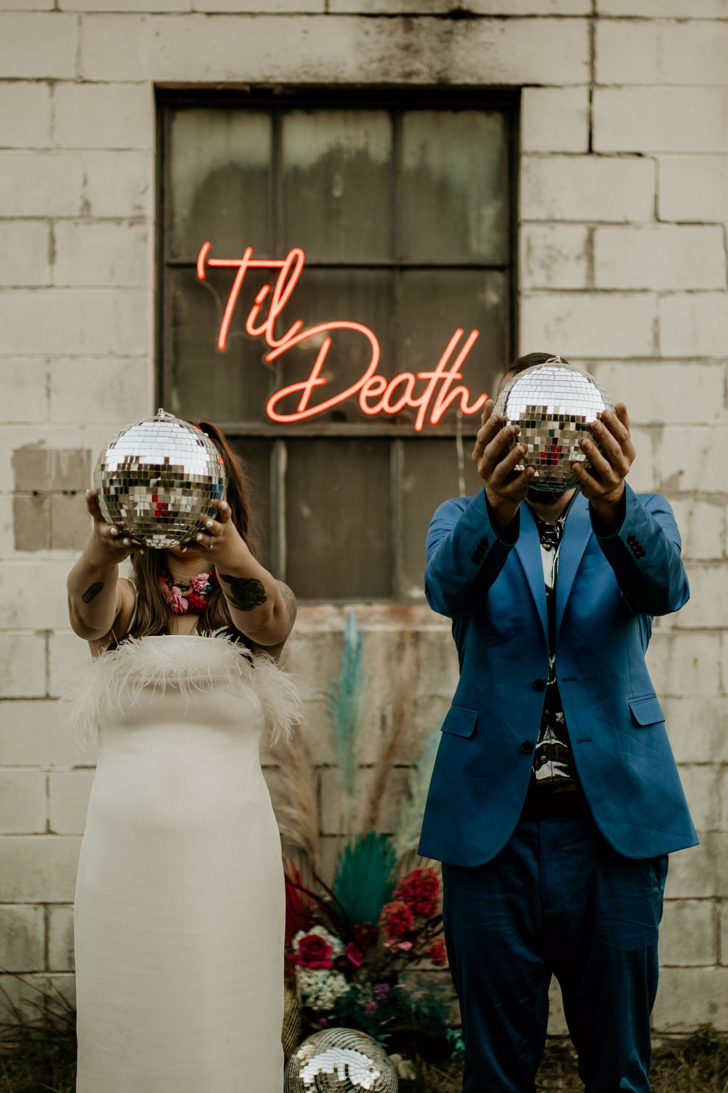 Couple holds disco balls in front of their faces before a glowing 'Til Death neon sign at an edgy wedding.