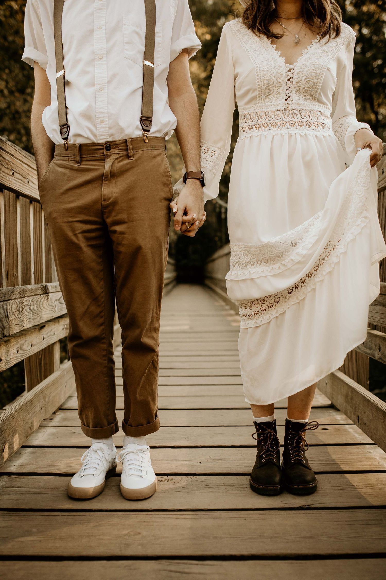 Couple holds hands on a wooden bridge, wearing casual boho wedding attire with suspenders and lace dress.