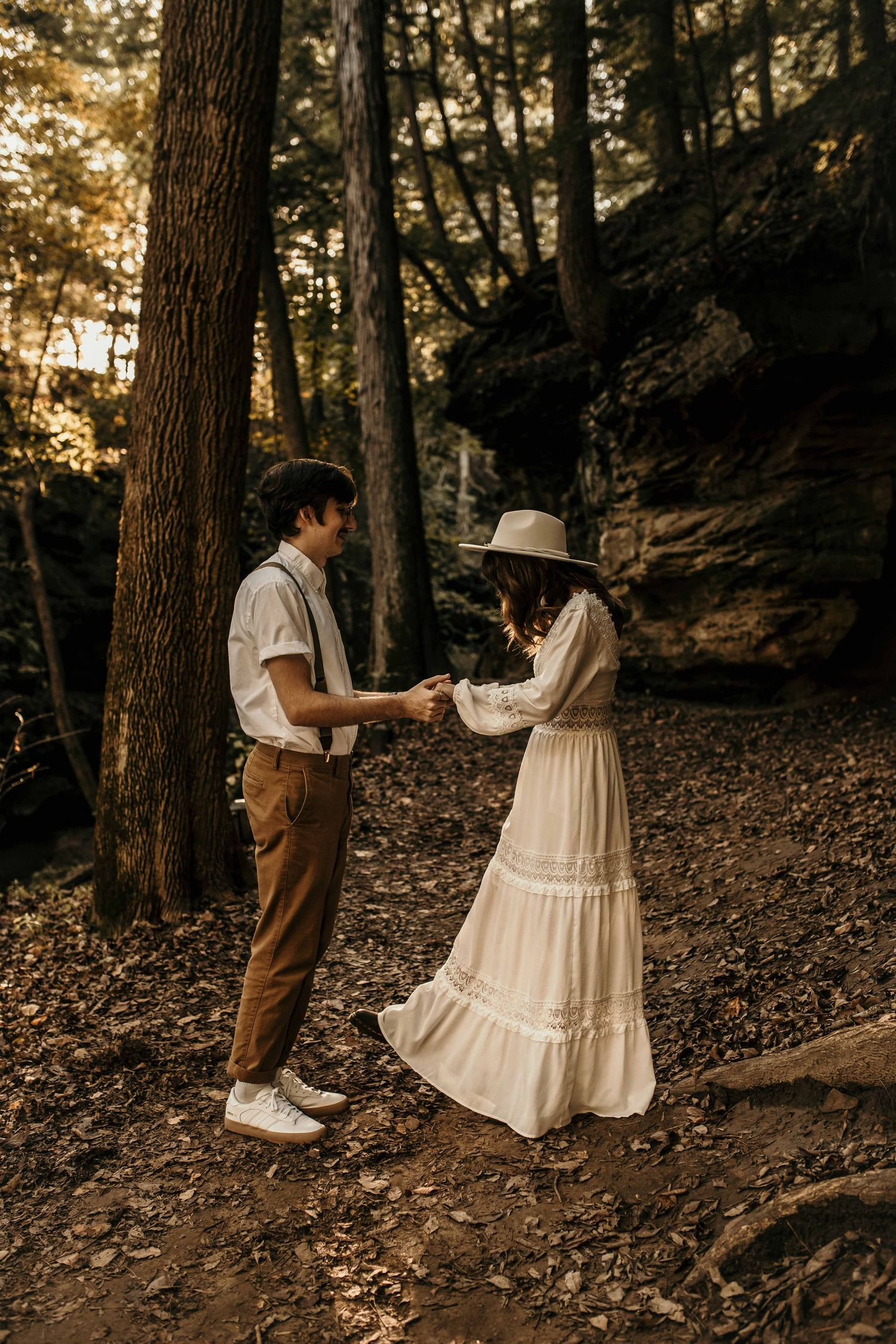 Couple holding hands in a sunlit forest, woman in white boho dress and hat, man in suspenders.