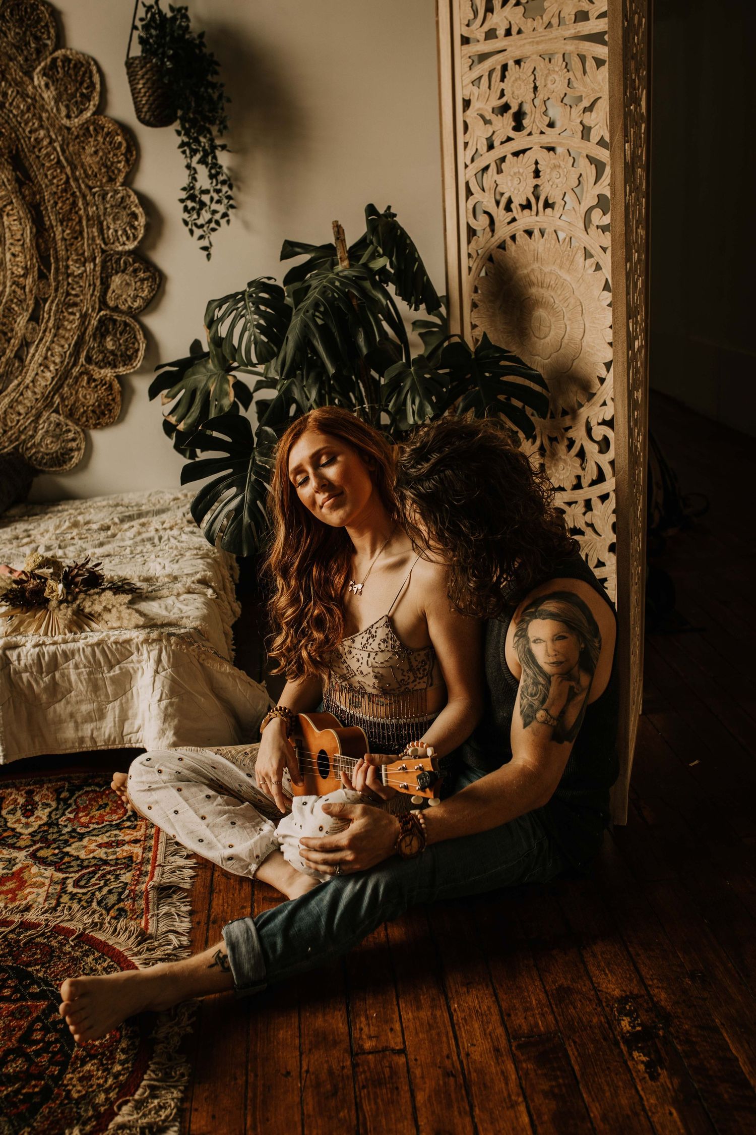 Bohemian woman with red hair sits on a wooden floor playing ukulele surrounded by eclectic boho decor.