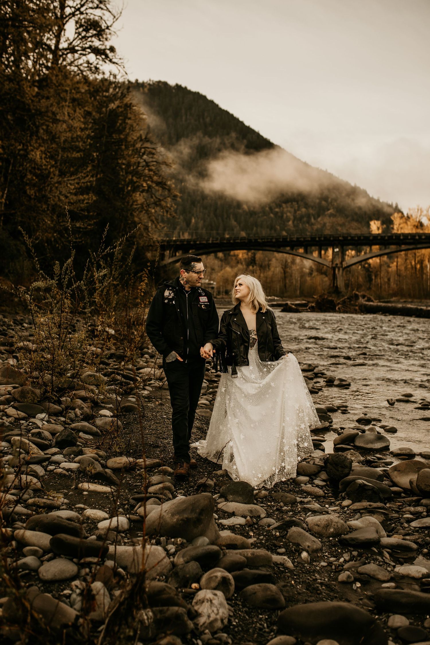 Bride in white dress and groom in leather jacket walking along a rocky riverside.