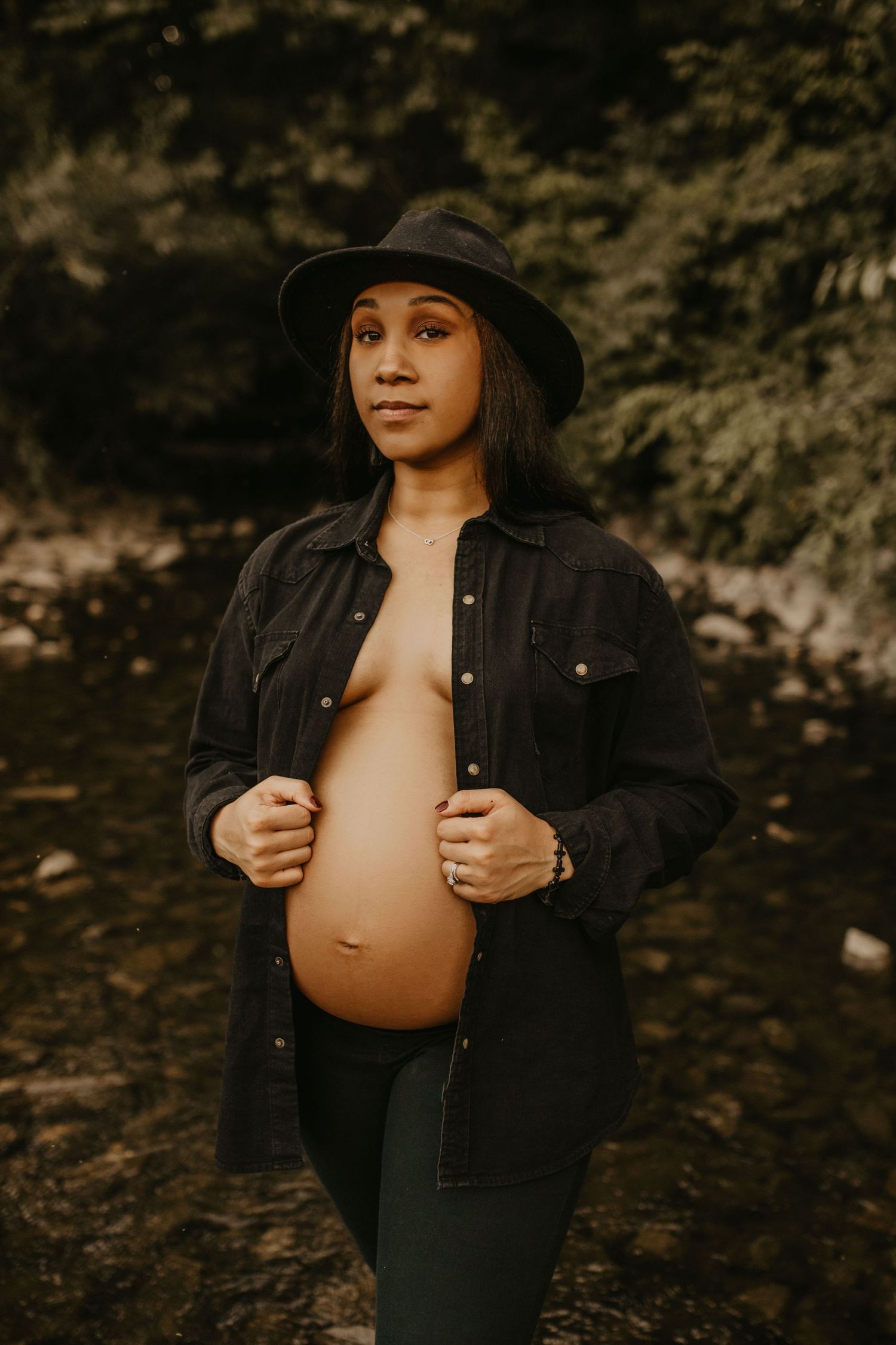Pregnant woman in black hat and denim jacket proudly displays her baby bump near a forest creek.