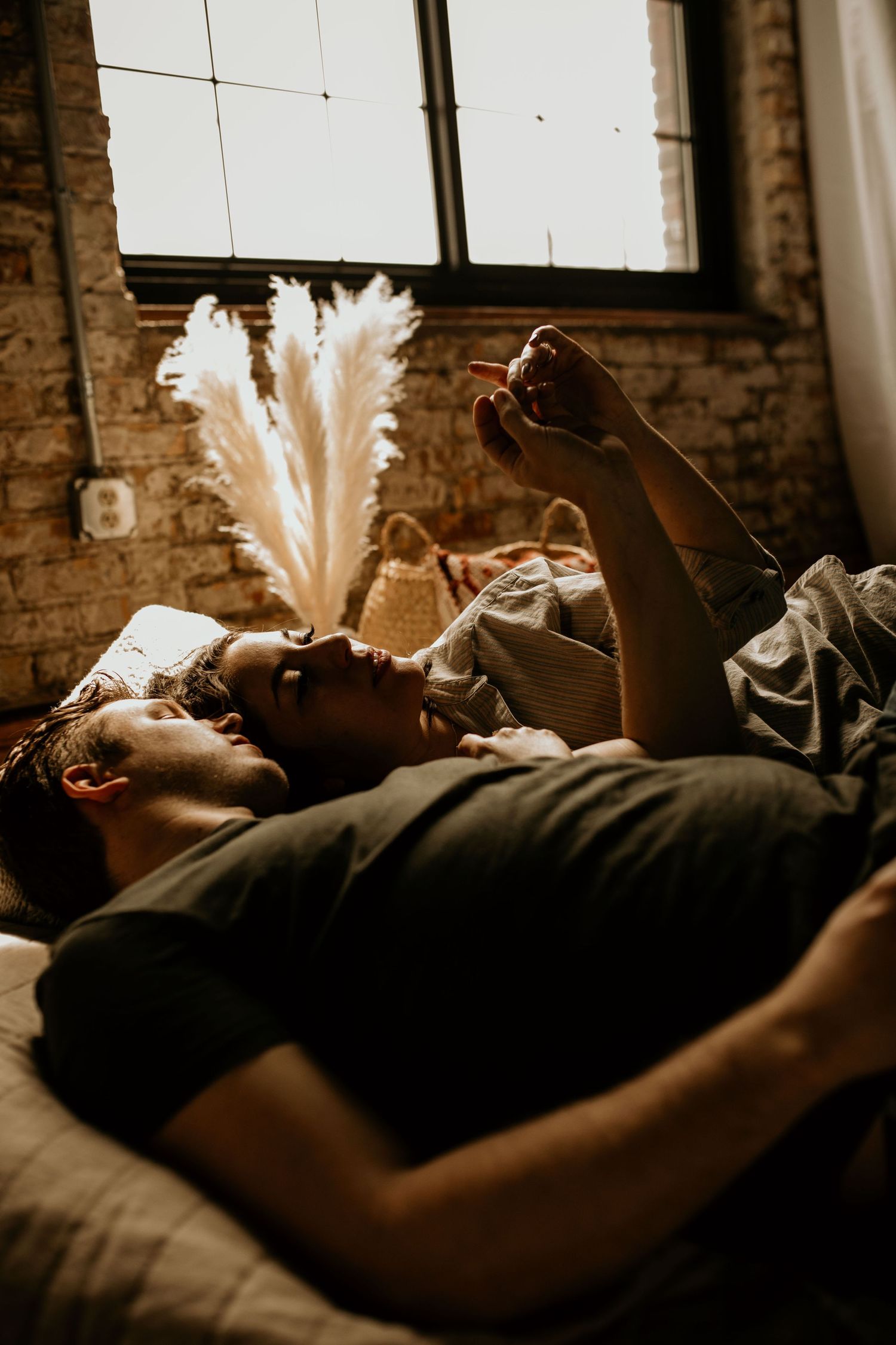 Romantic couple lies on a bed in a sunlit loft apartment with exposed brick and pampas grass decor.