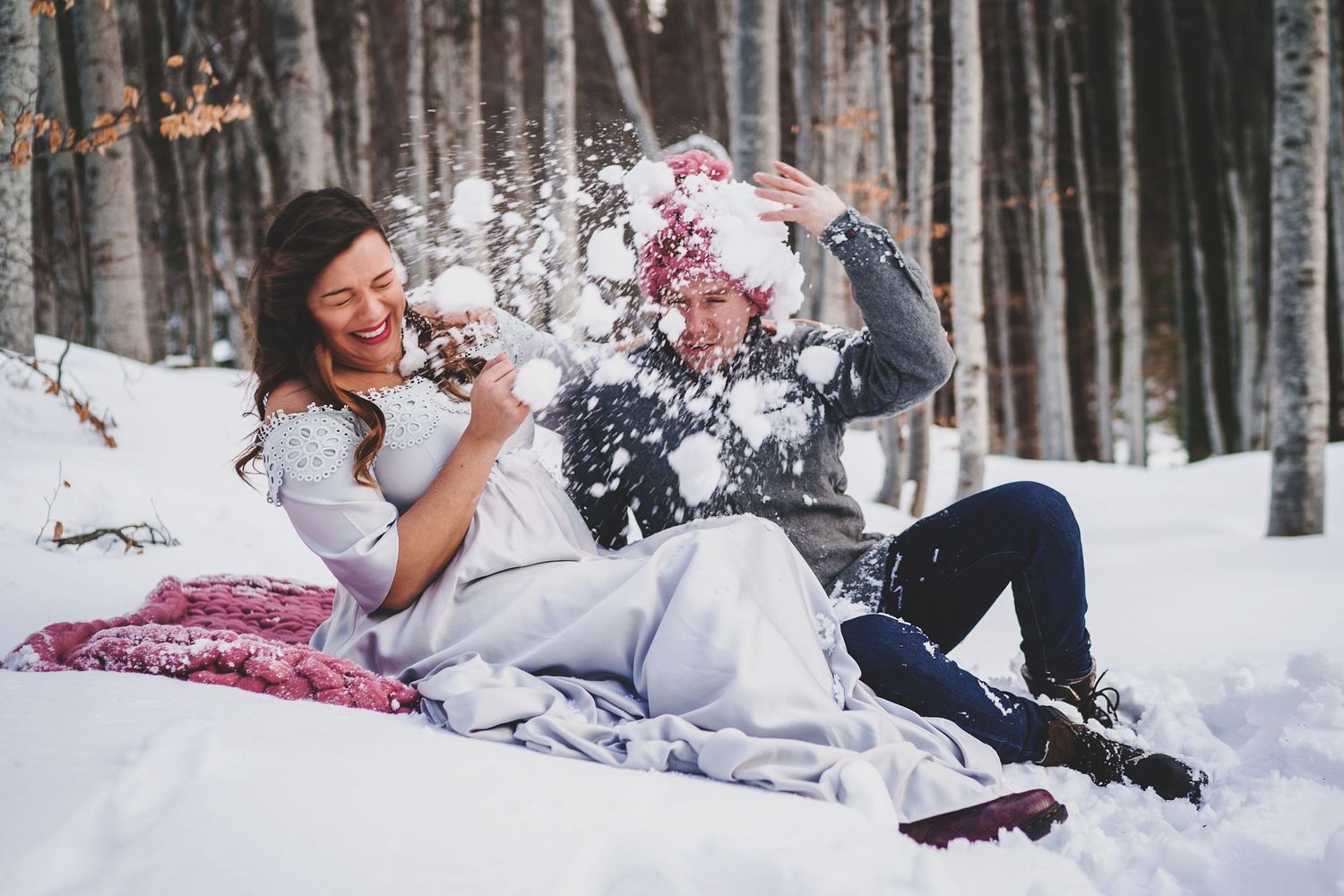 Playful winter photoshoot with couple throwing snow while lying on white ground.