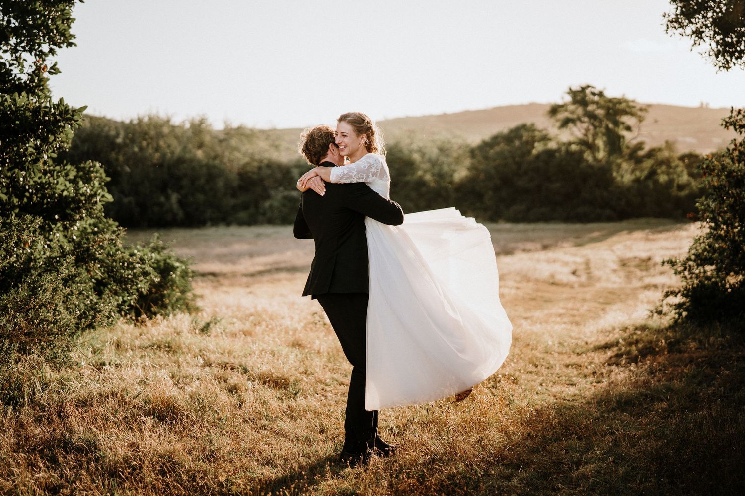 Romantic wedding portrait with groom lifting bride against warm sunset light.