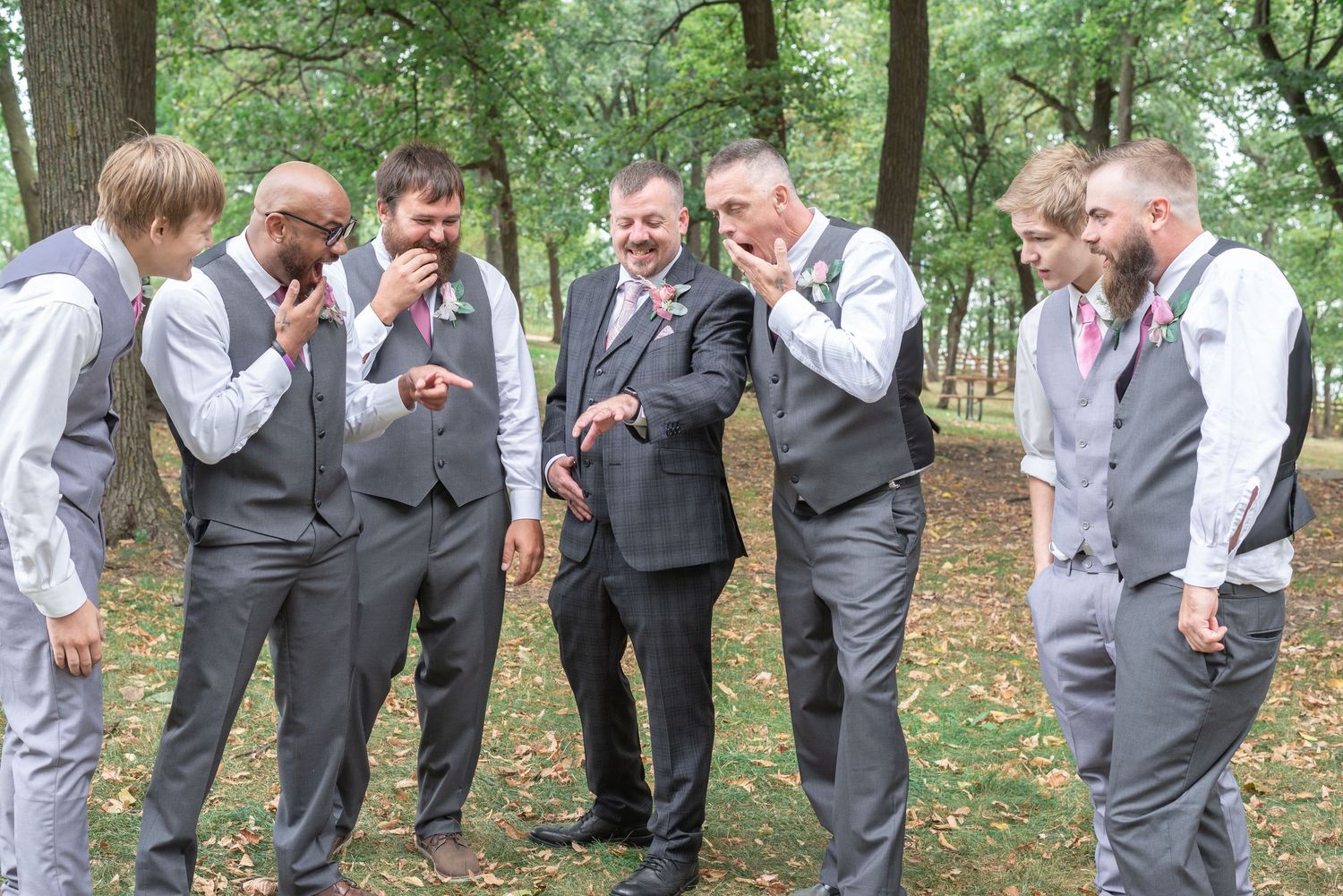 Groom surrounded by his groomsmen in the woods as they all fawn over his wedding band for funny picture